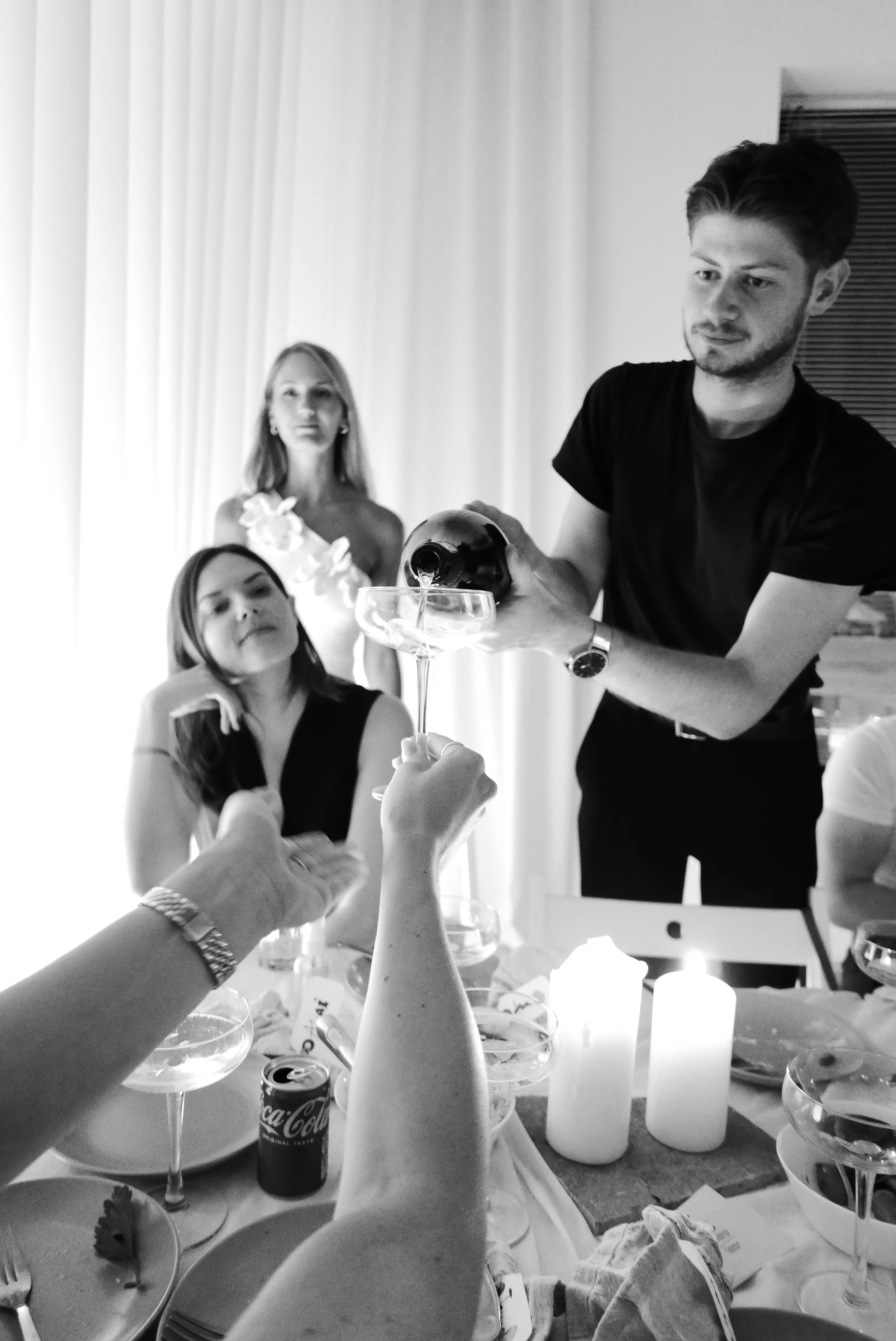 People celebrating with drinks at a dinner table, with one person pouring champagne into a glass, in a black-and-white photo.