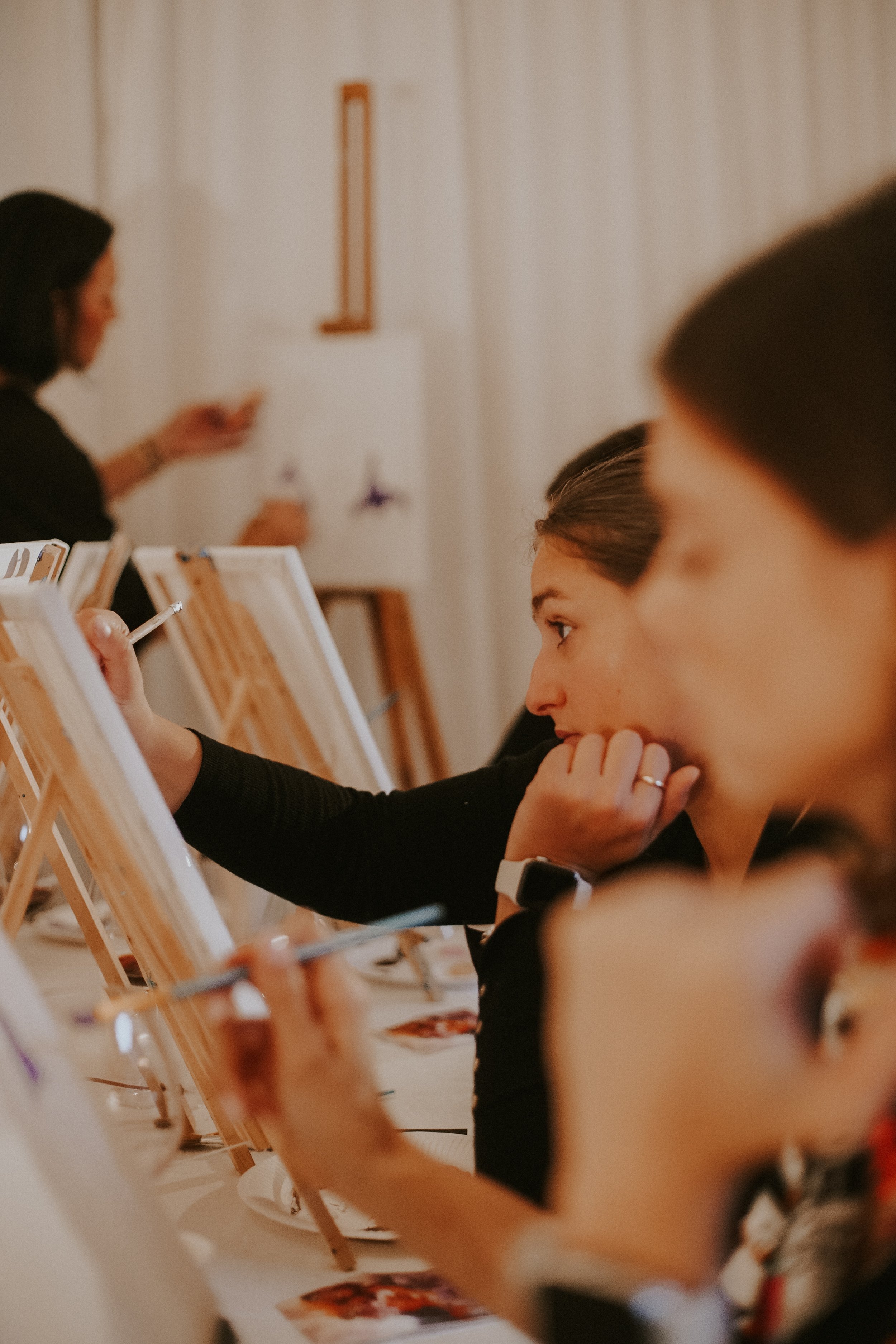 People participating in a painting class, focusing on their canvases and painting on small easels.