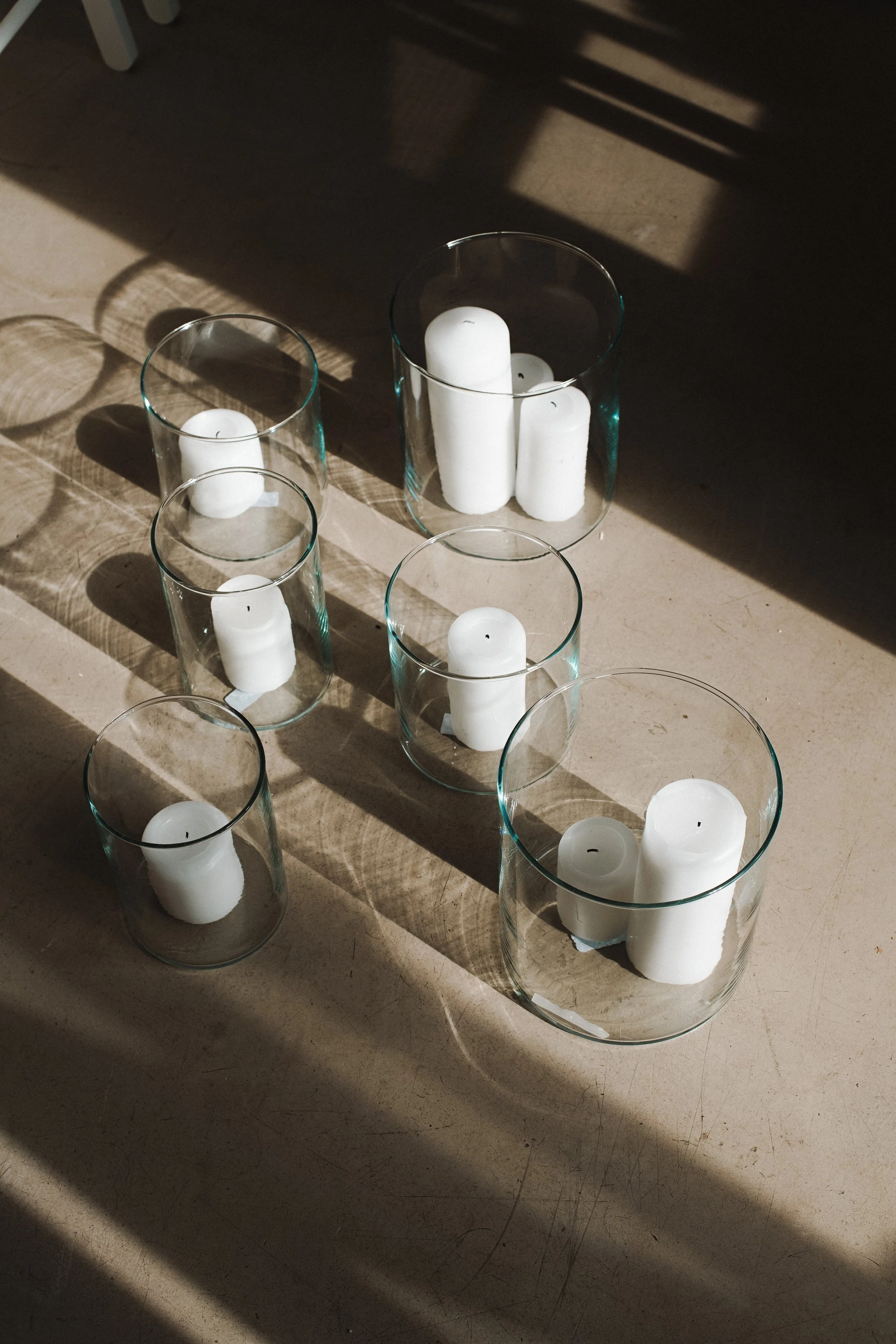 Six glass vases with white candles inside, placed on a beige surface, with shadows cast by sunlight and a patterned shadow from an unseen object.
