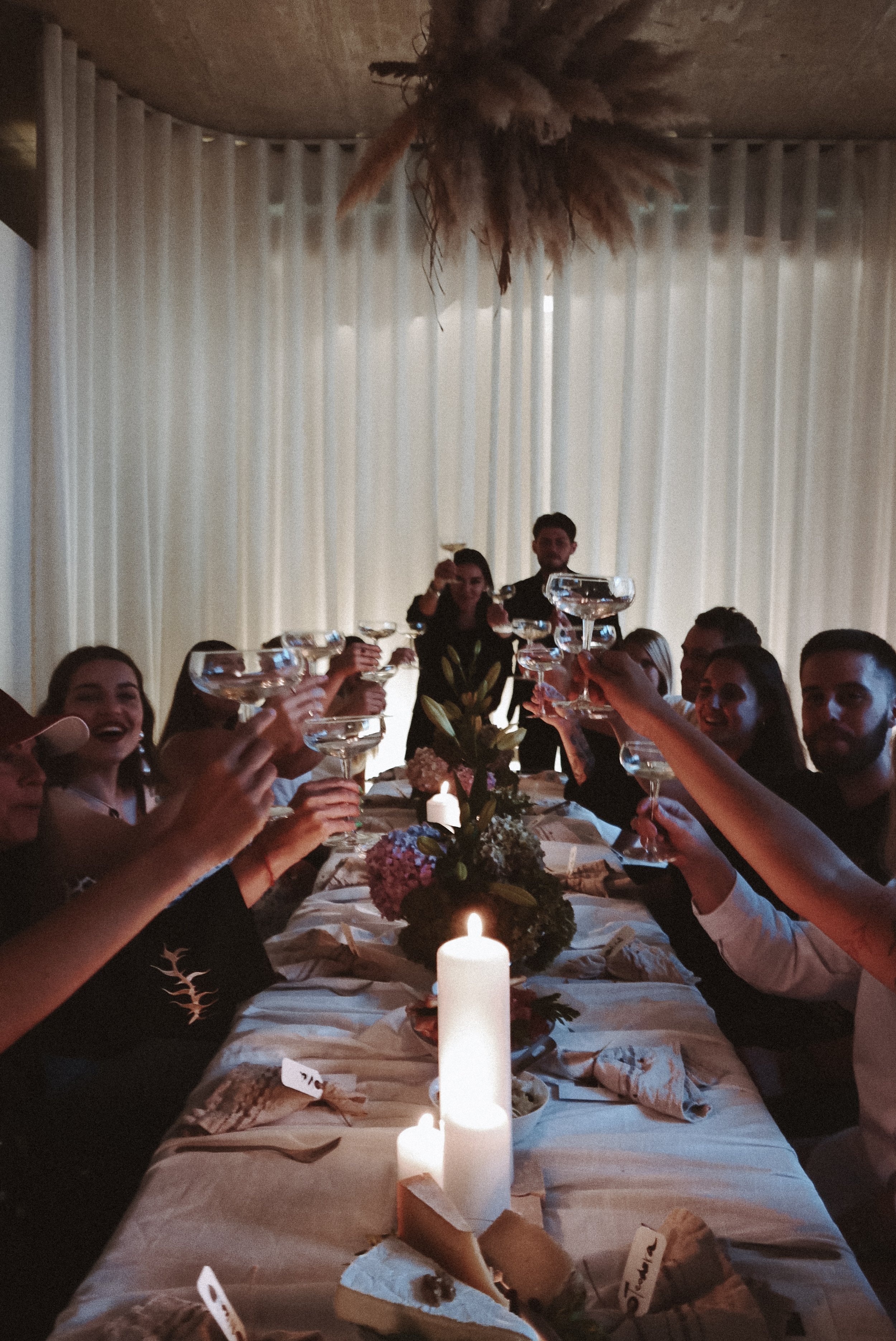 People celebrating at a dinner party raising glasses in a toast, with candles and a floral centerpiece on the table, and sheer curtains in the background.