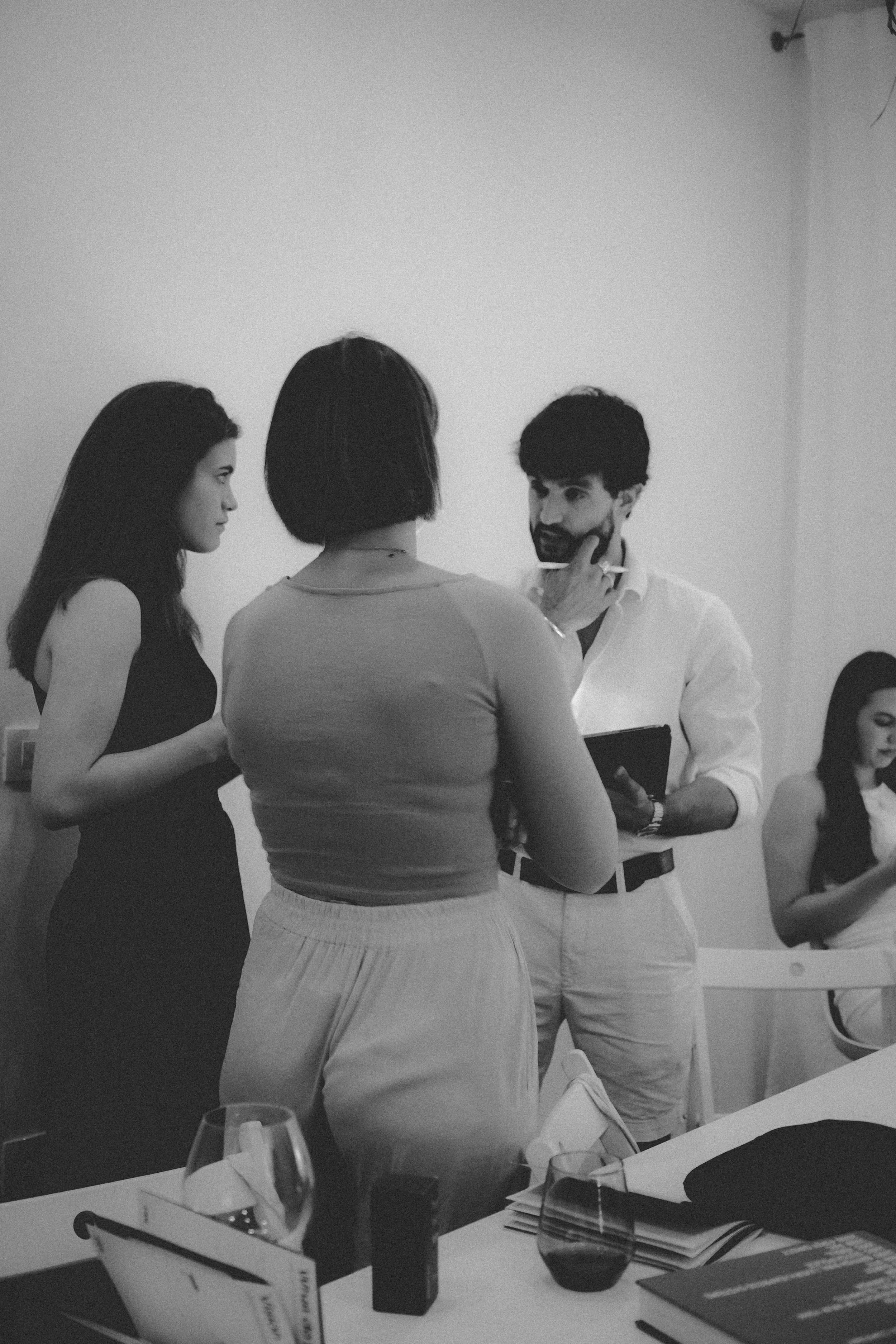 Group of people having a conversation in a meeting room, with one woman touching the man's face, two women sitting in the background, and a table with glasses, papers, and a book.