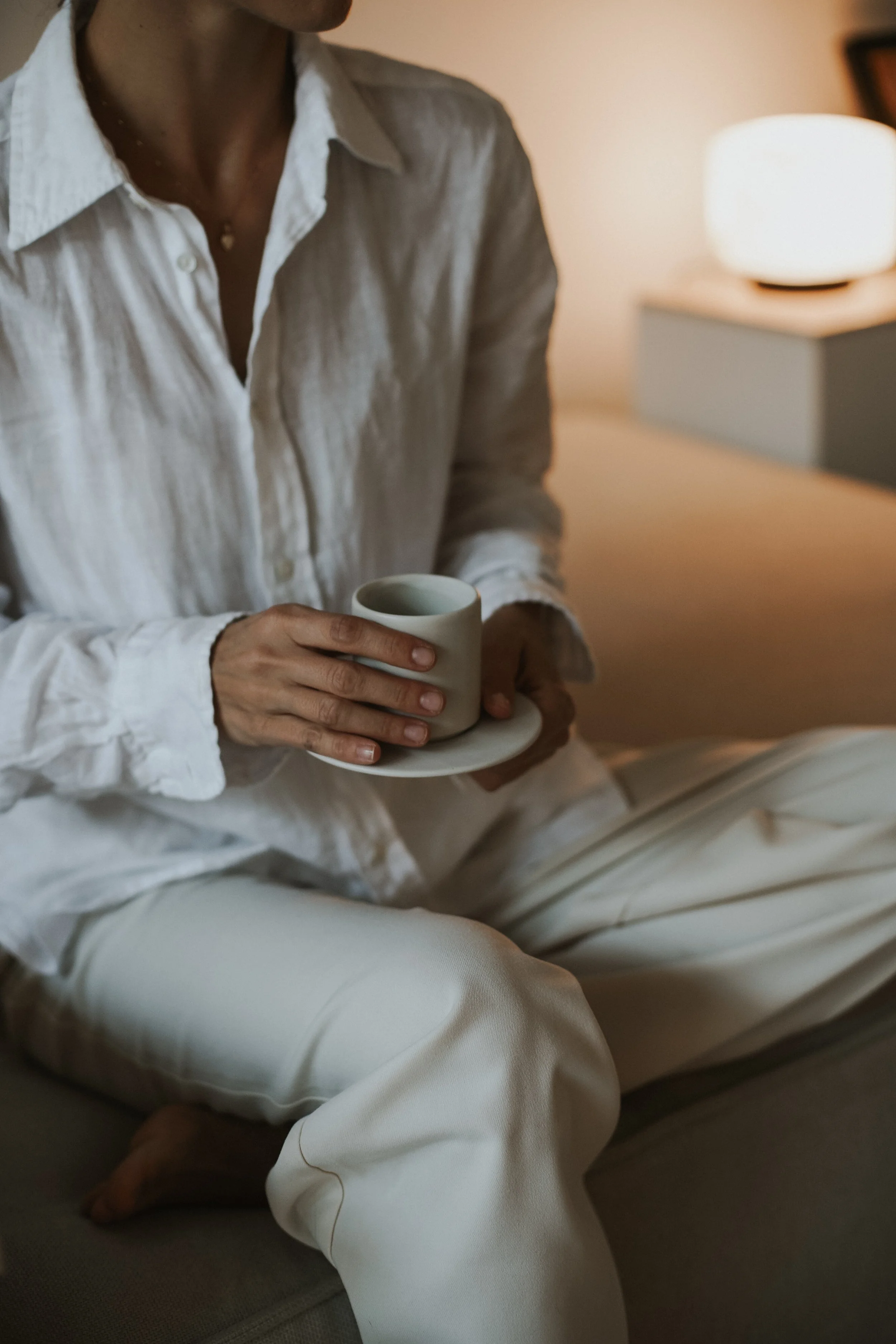Person sitting on a couch, wearing a white shirt and cream-colored pants, holding a cup of coffee or tea in a small plate, in a cozy, softly lit room with a nightstand and a table lamp.