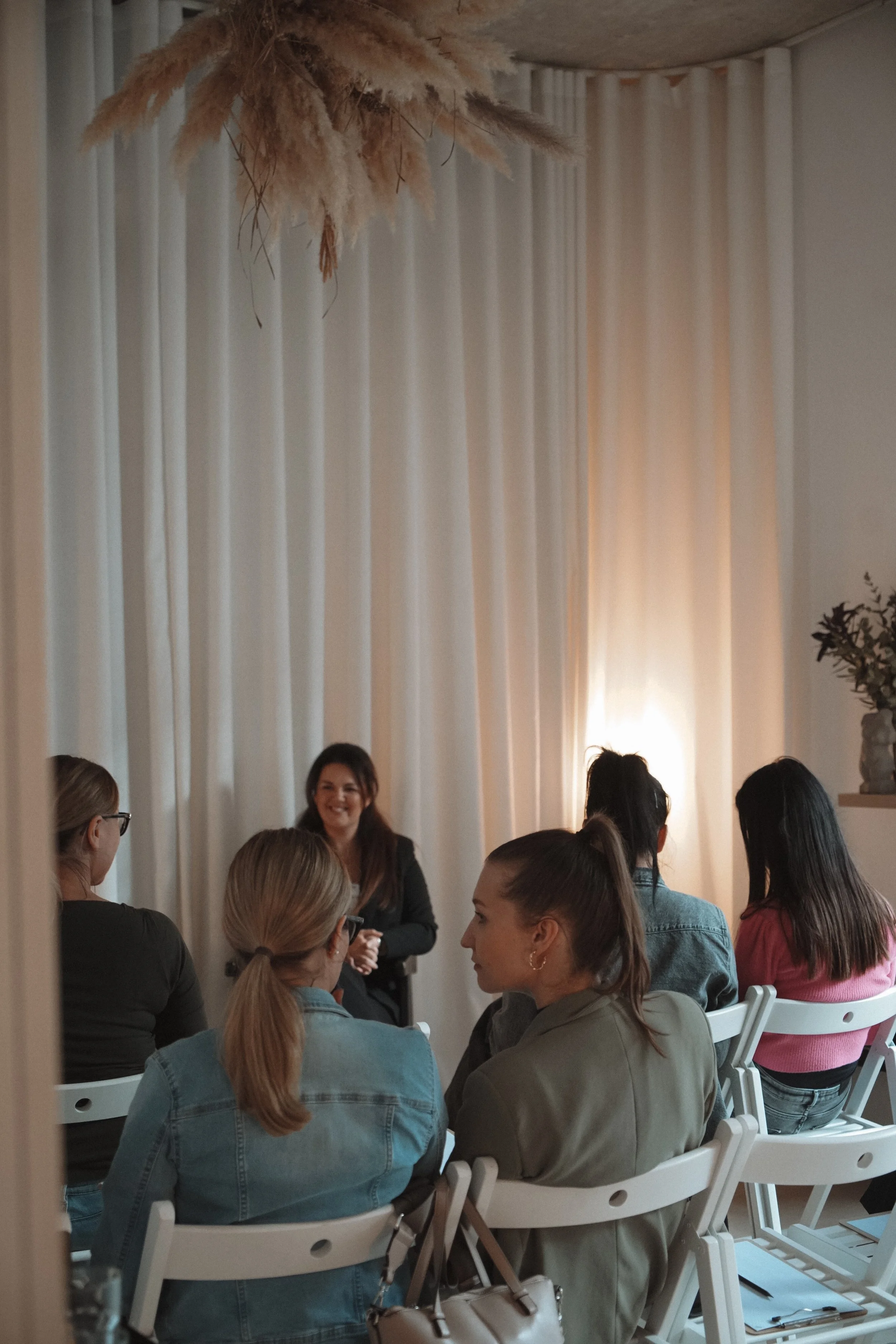 A woman is speaking or presenting to a group of women seated in a room with white curtains and soft lighting.