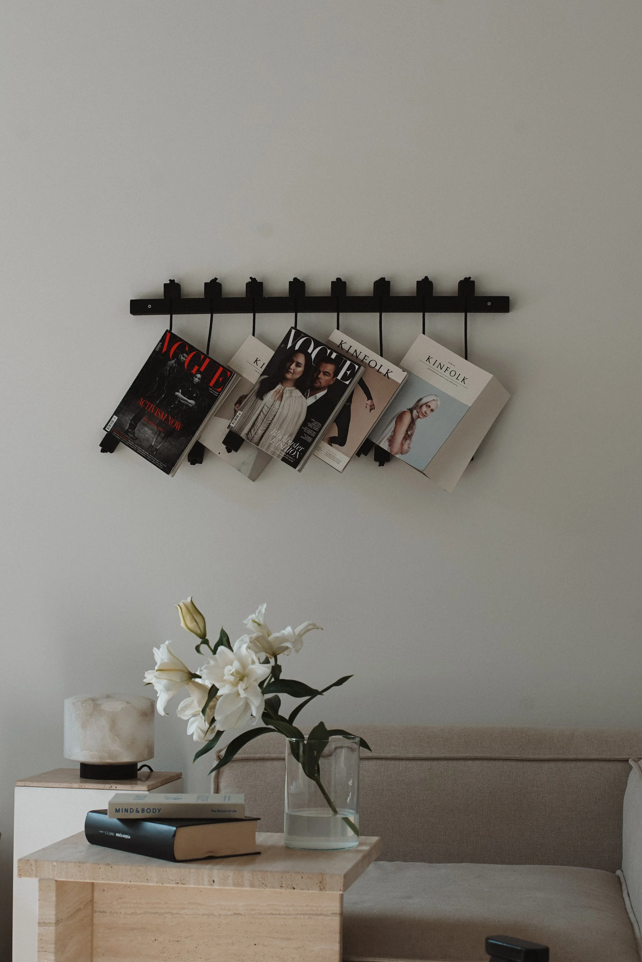 A living room with a beige sofa, a wooden coffee table holding a glass vase with white lilies and two books, and a white table lamp on a white side table. On the wall, a black wall-mounted magazine rack with five Kate magazines.