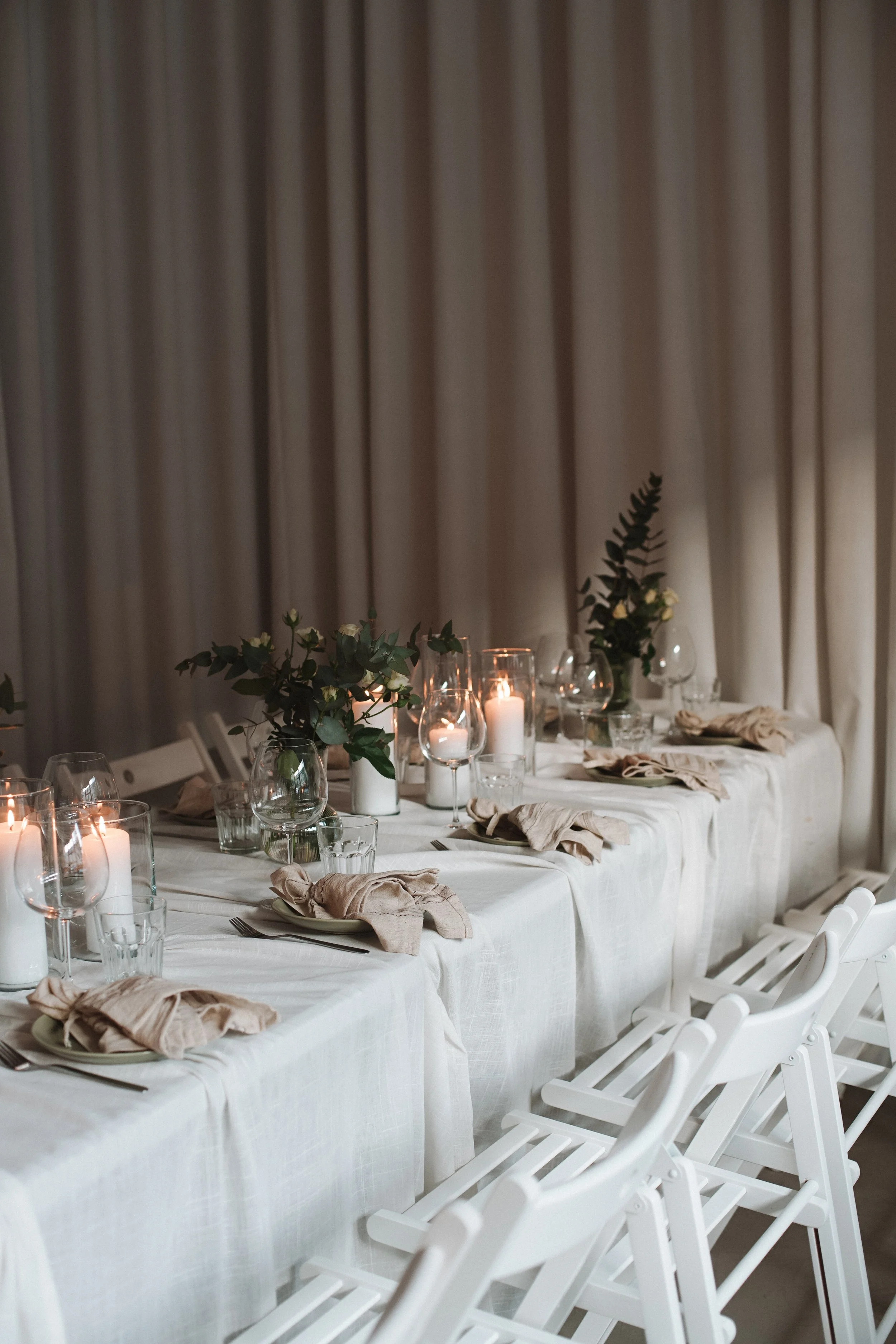 A decorated dining table with white tablecloth, beige napkins, glassware, candles, and green foliage arrangements, set in front of beige curtains.