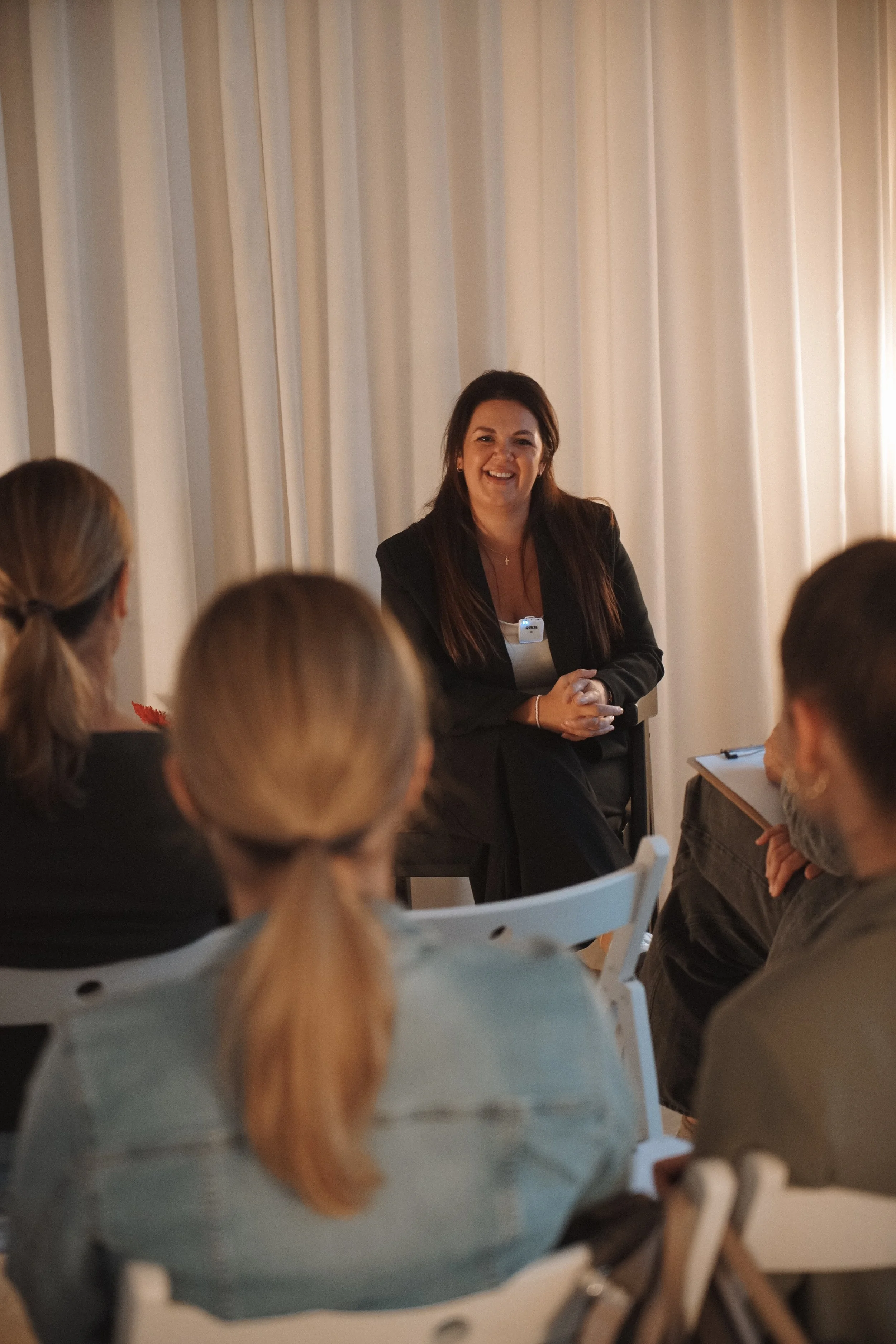 A woman in a black blazer speaking to a group of people in a room with curtains, with her hands clasped and a microphone clipped on her blazer.
