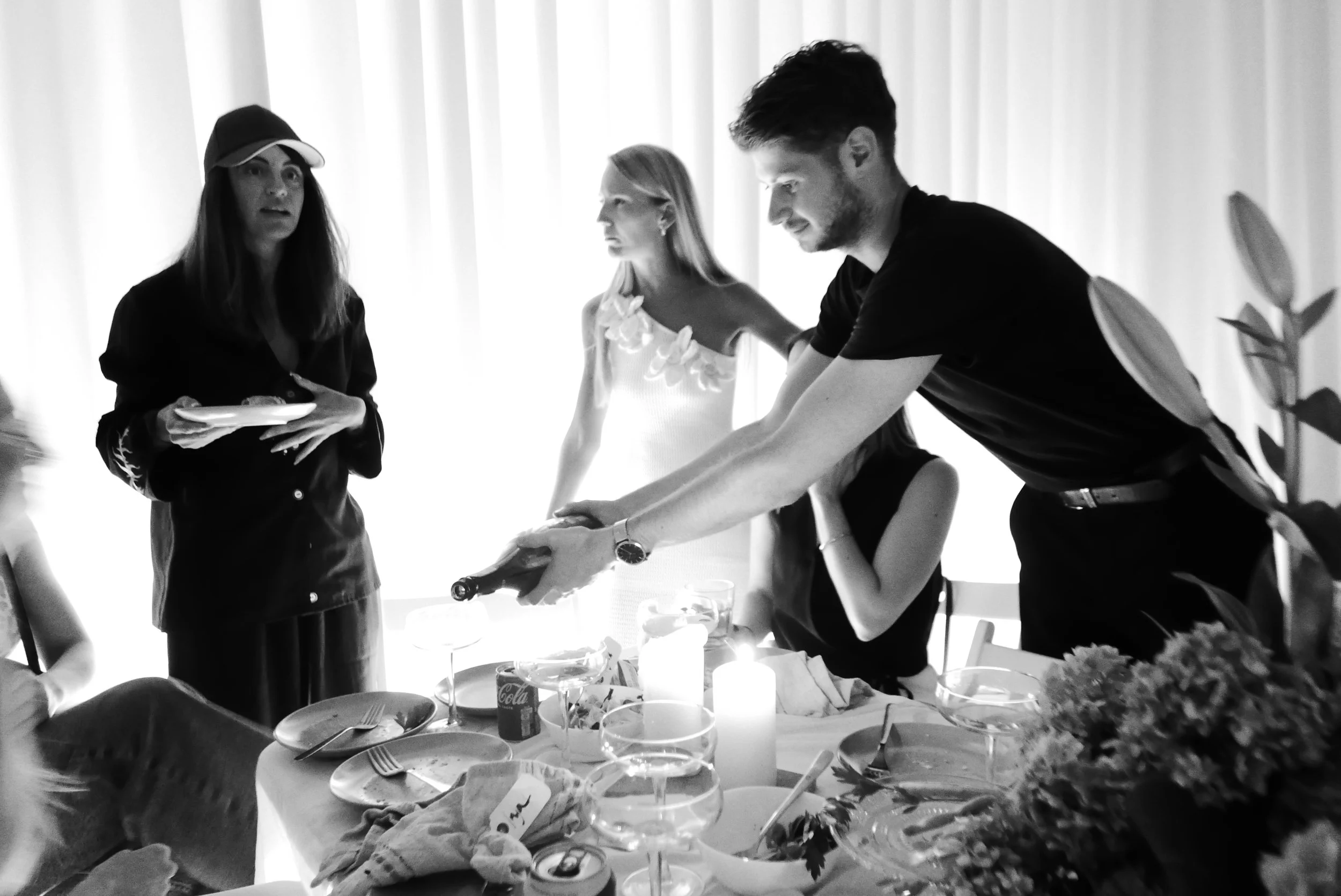 A group of people gathered around a dining table with candles and flowers. One person is serving wine, while another woman holds a plate, and others are seated or standing nearby.