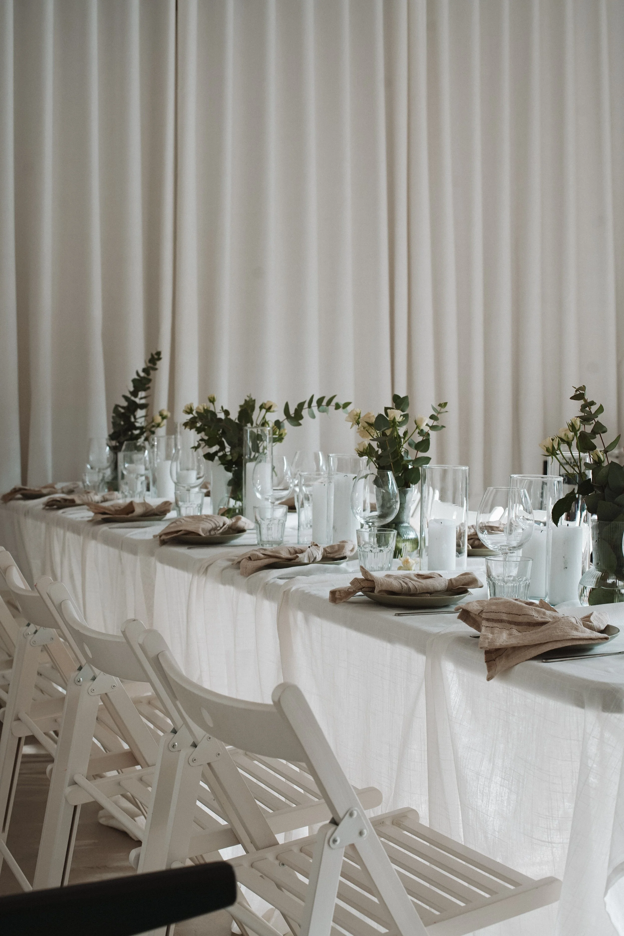Long banquet table decorated with glassware, napkins, and floral centerpieces, set against a backdrop of white curtains.
