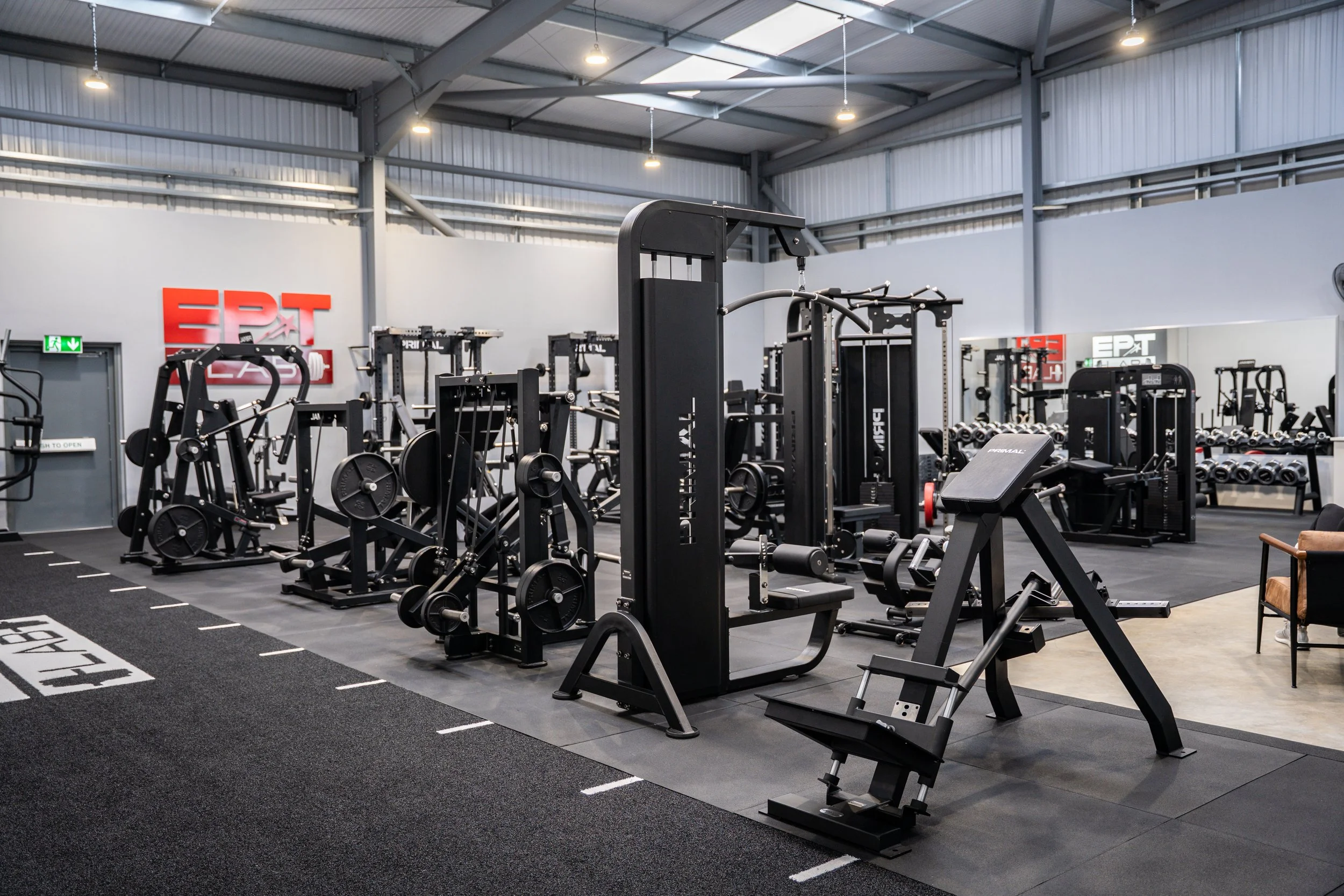 Inside a modern gym with various black exercise equipment, including weight machines and free weights, in a spacious, well-lit area with a grey ceiling and walls, and a red sign reading ЕРАТ on the wall.
