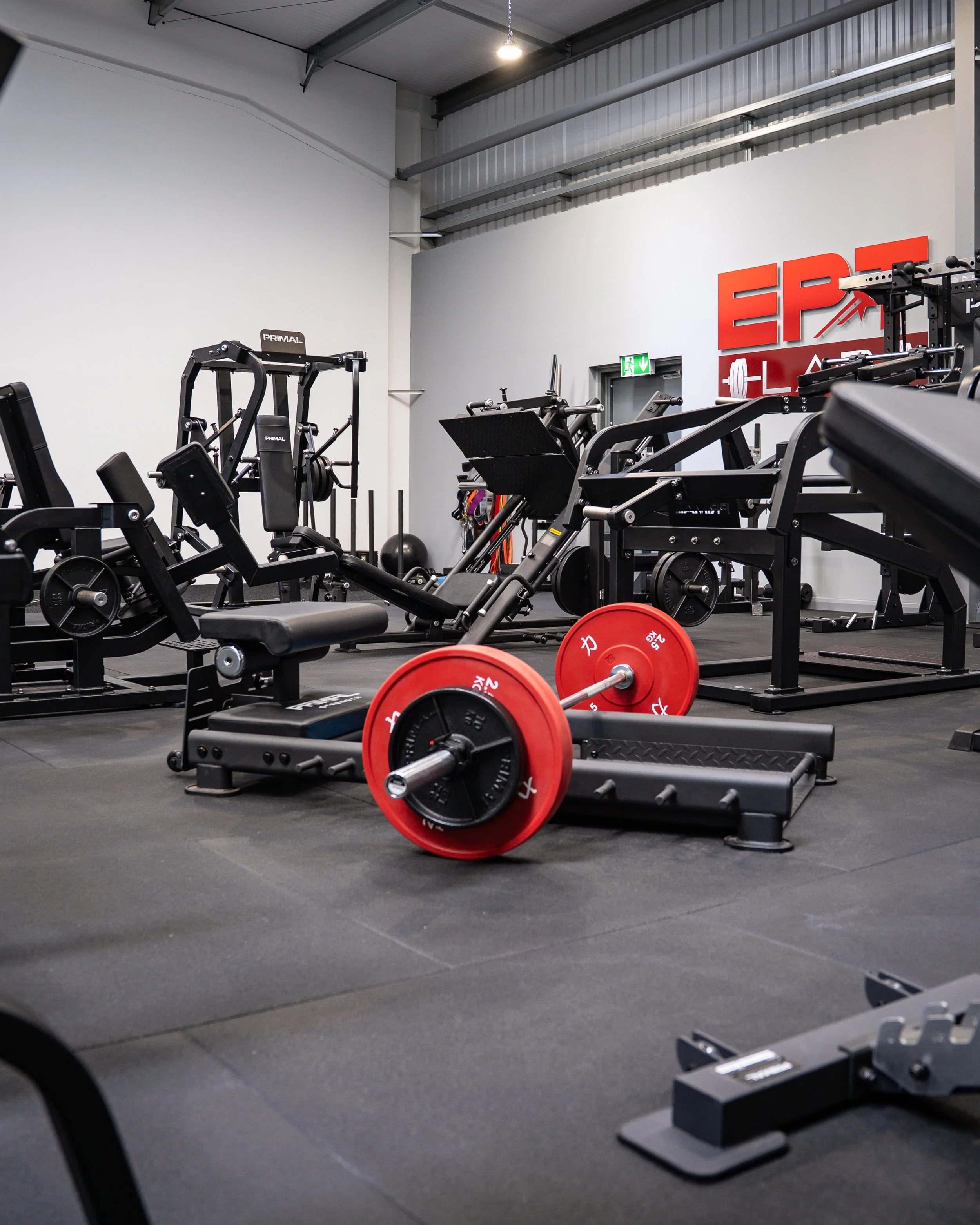 Interior of a gym with various exercise equipment, including a barbell with red weight plates lying on the floor, adjustable benches, weight machines, and a black exercise bike, all set on black rubber flooring. There is a large red sign on the wall reading 'EPIC' in the background.
