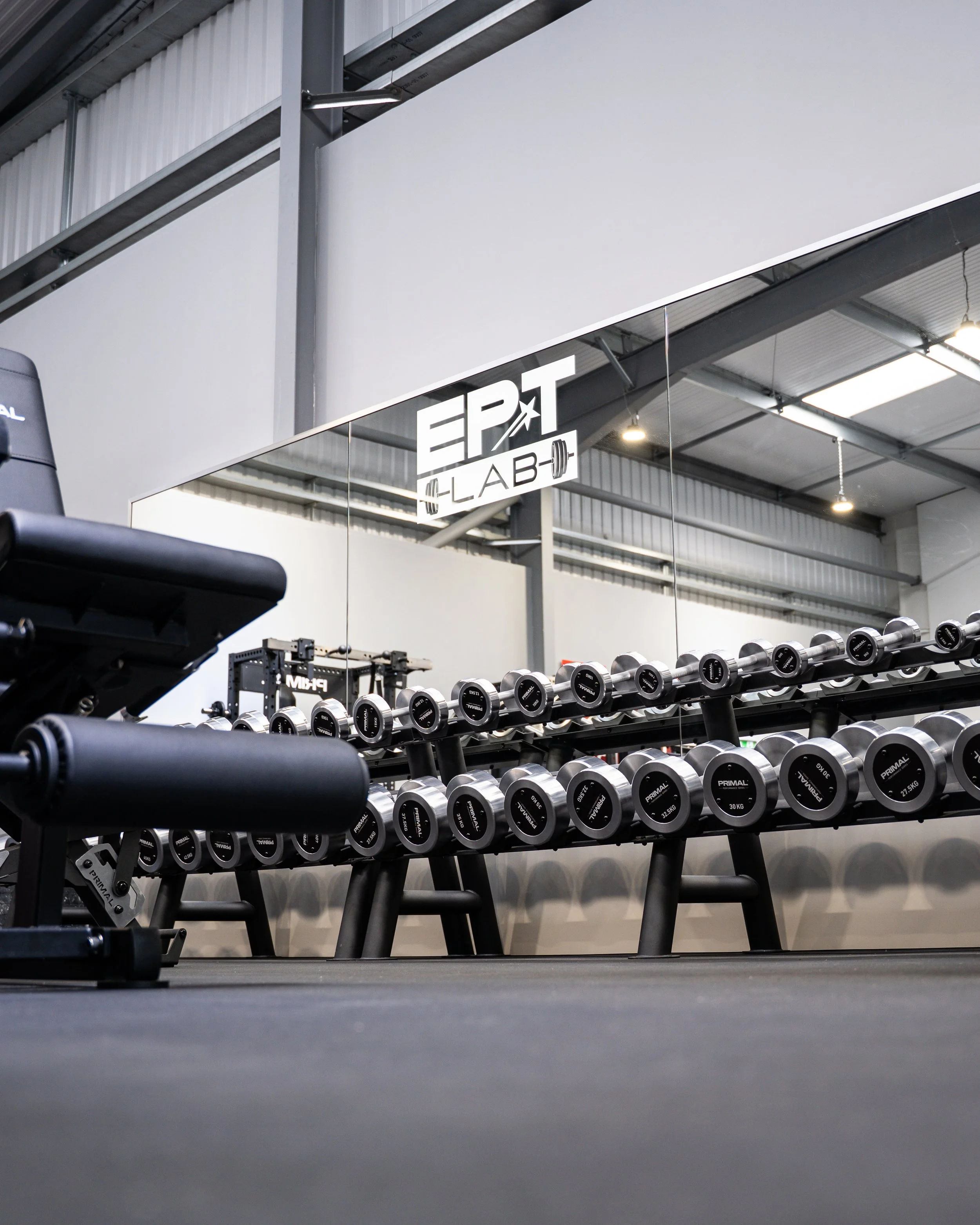 Dumbbells arranged on a black rack in a gym, with a mirror and a sign that reads 'EPT LAB' in the background.