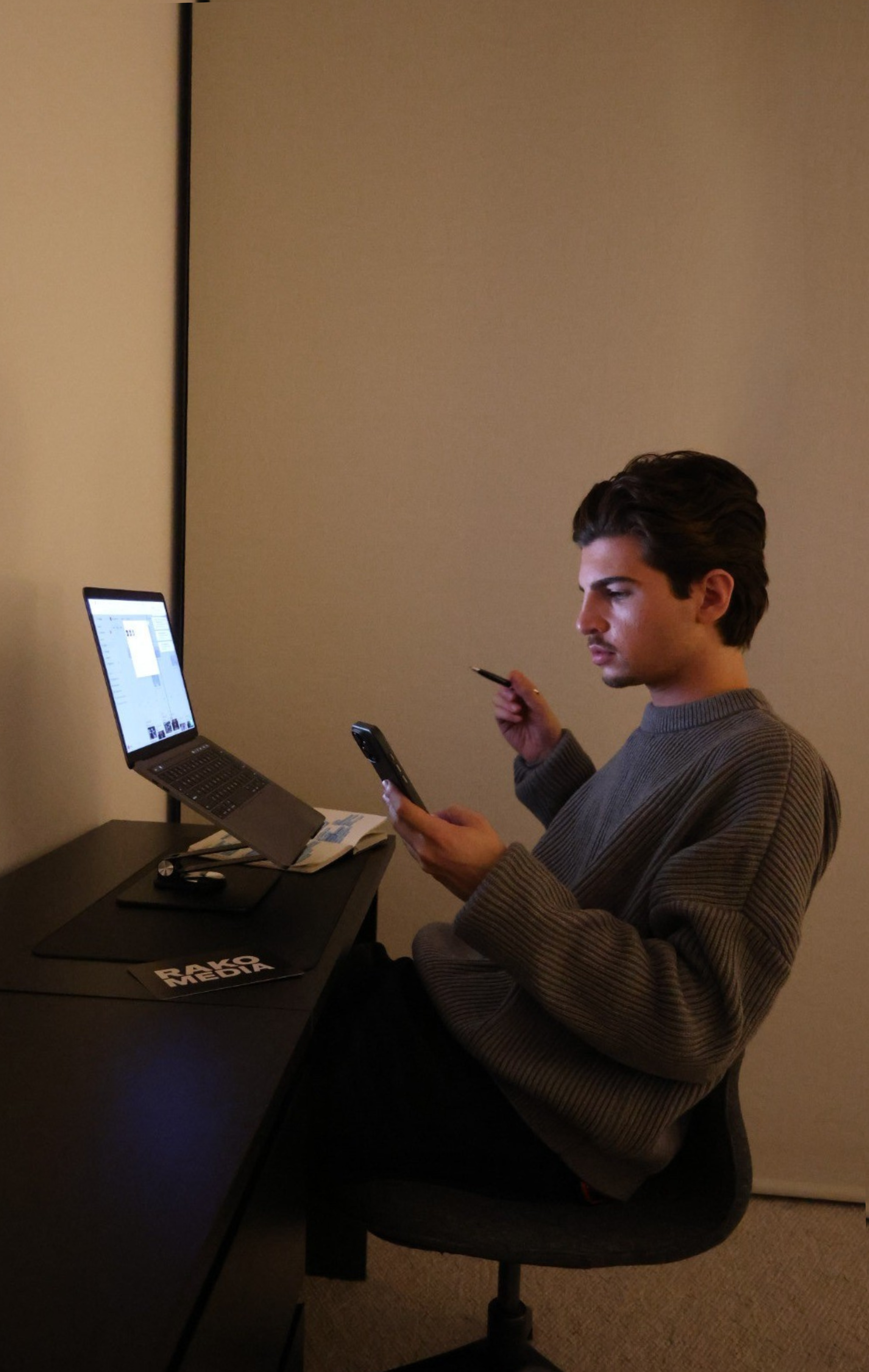 Young man sitting at a desk, looking at his phone while holding a pen, with a laptop in front of him and a newspaper titled 'RAKO MEDIA' beside it, in a dimly lit room.