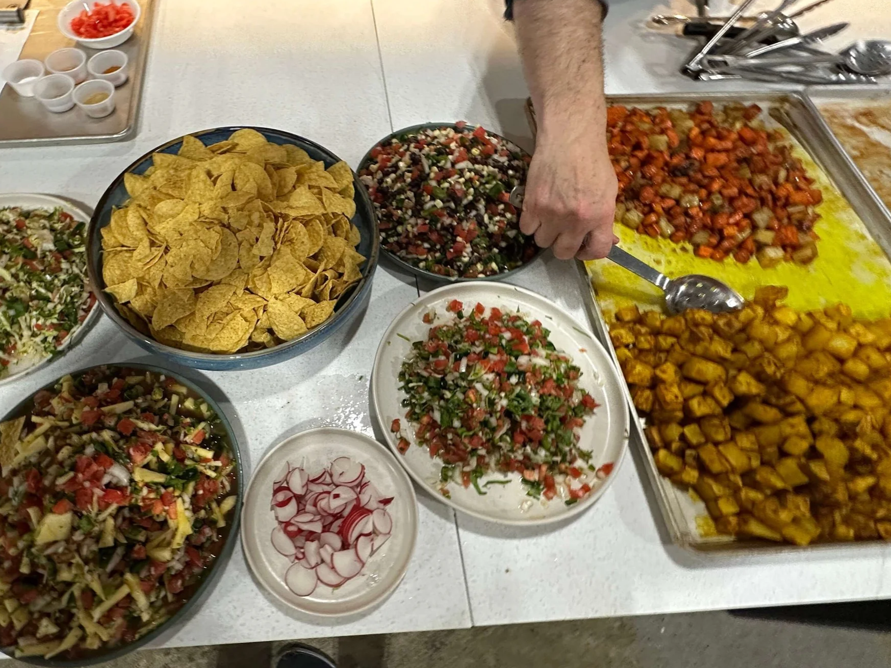 An assortment of Mexican foods including tortilla chips, two different kinds of salsas, chopped radishes, chopped tomatoes, and cooked diced sweet potatoes on a table.