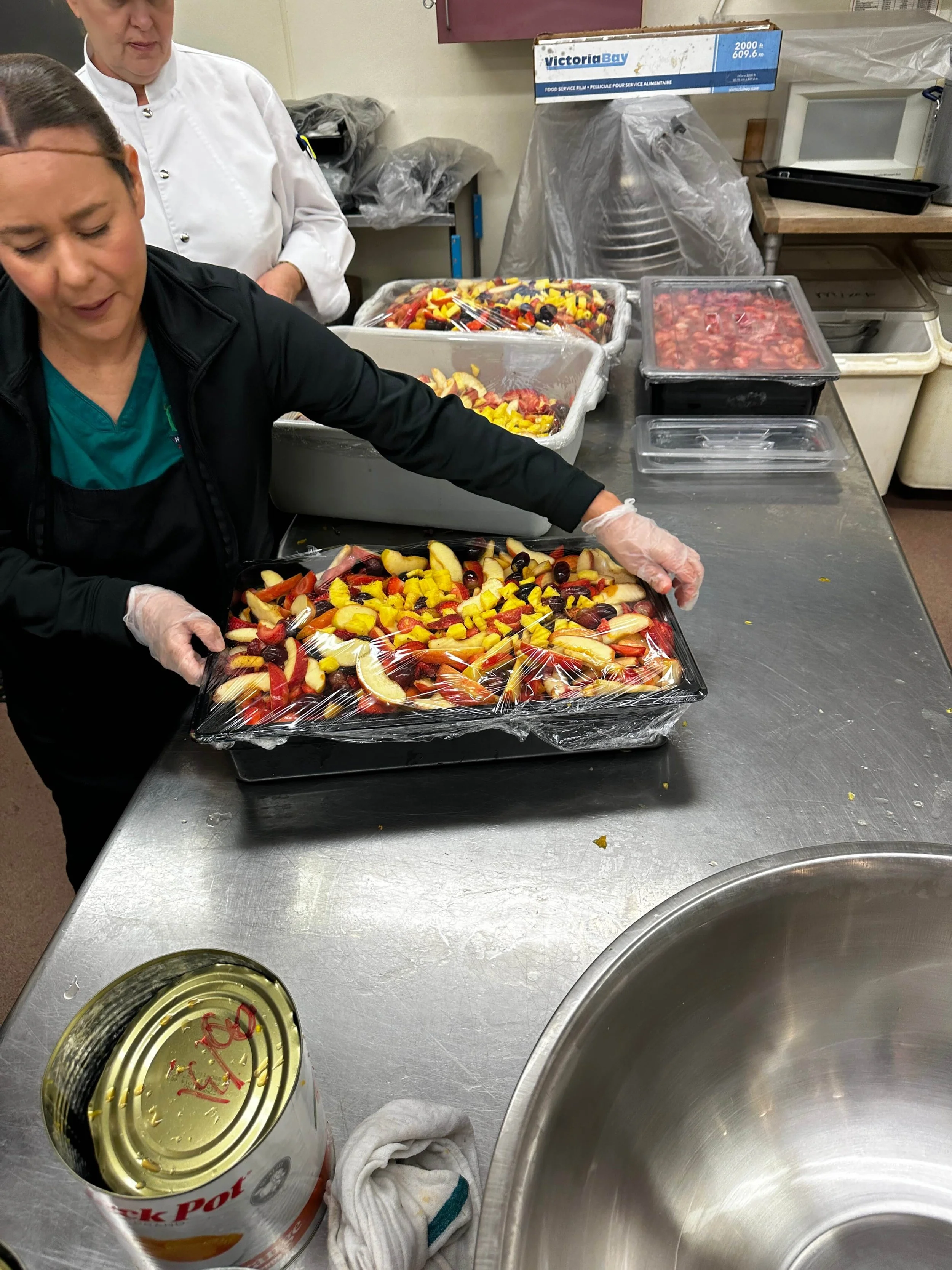Workers preparing or packing canned and bottled food items, with a focus on an open canned item in a stainless steel container and a large tray of sliced fruit or salad in the background.