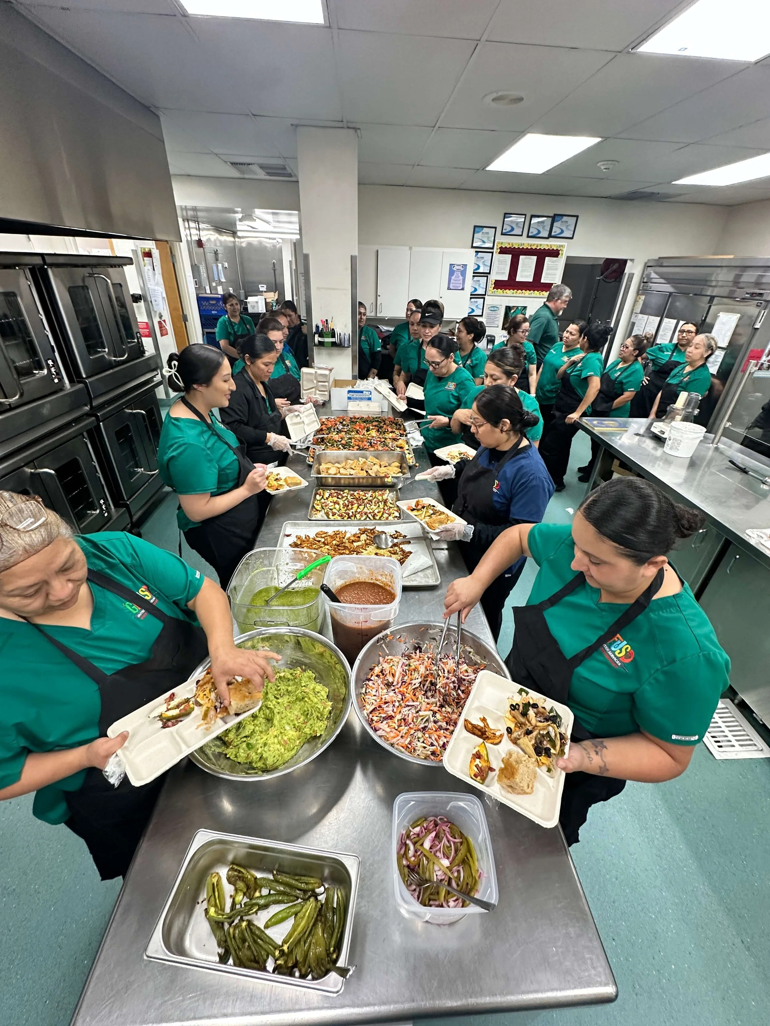 A group of people in green uniforms serve themselves food at a cafeteria counter with various dishes, including salads, pickles, and cooked vegetables.
