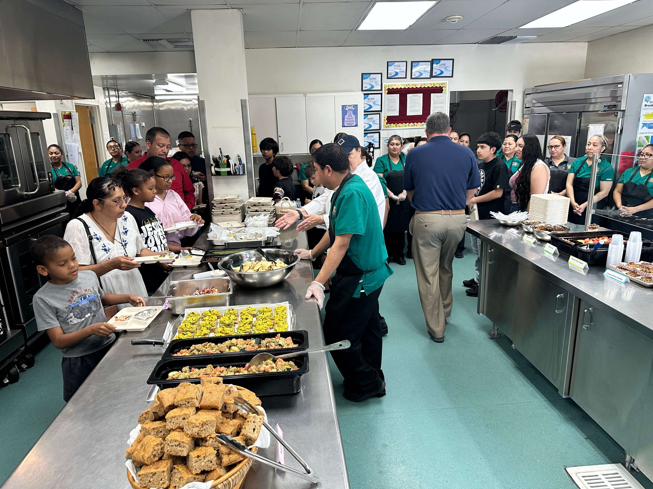 Food service line with various dishes, salad, and baked goods, and people waiting in line in a cafeteria.
