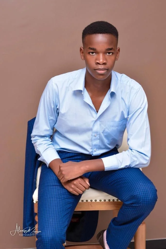 Young man with short hair and serious expression, sitting on a chair against a plain beige background, wearing a light blue dress shirt and dark blue checkered pants.
