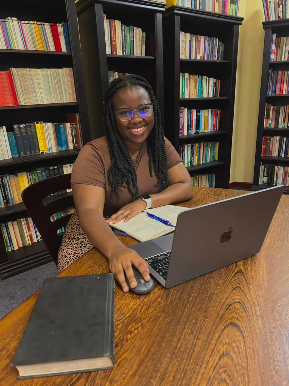 A smiling woman with glasses sitting at a wooden table in a library, using a laptop, with a notebook, pen, and a closed book on the table, surrounded by tall bookshelves filled with colorful books.