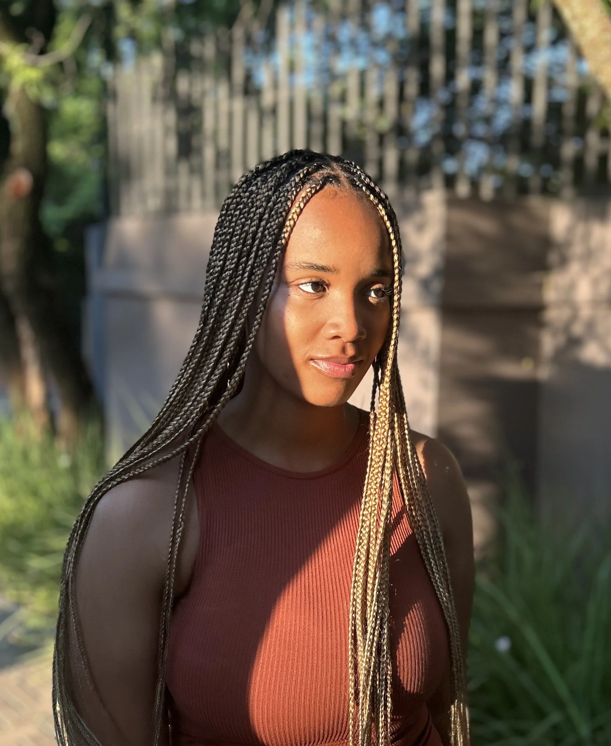 A woman with braided hair standing outdoors in sunlight, wearing a reddish-brown sleeveless top, with a blurred background of greenery and a fence.