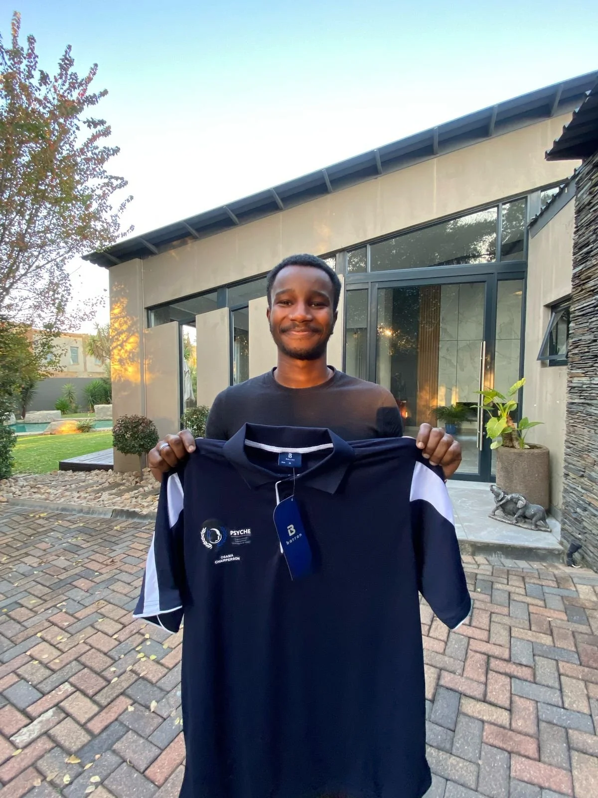A young man with short curly hair and a beard holding a navy blue sports shirt outside in a backyard with a modern house, trees, and a patio.