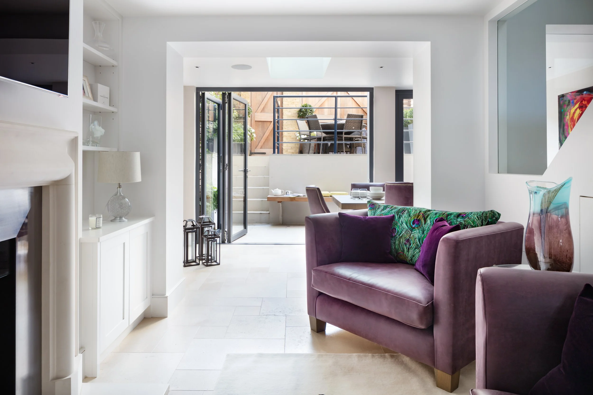 Living room with purple sofa, colorful pillows, and glass vases, opening to outdoor patio with dining table and chairs, bright natural lighting