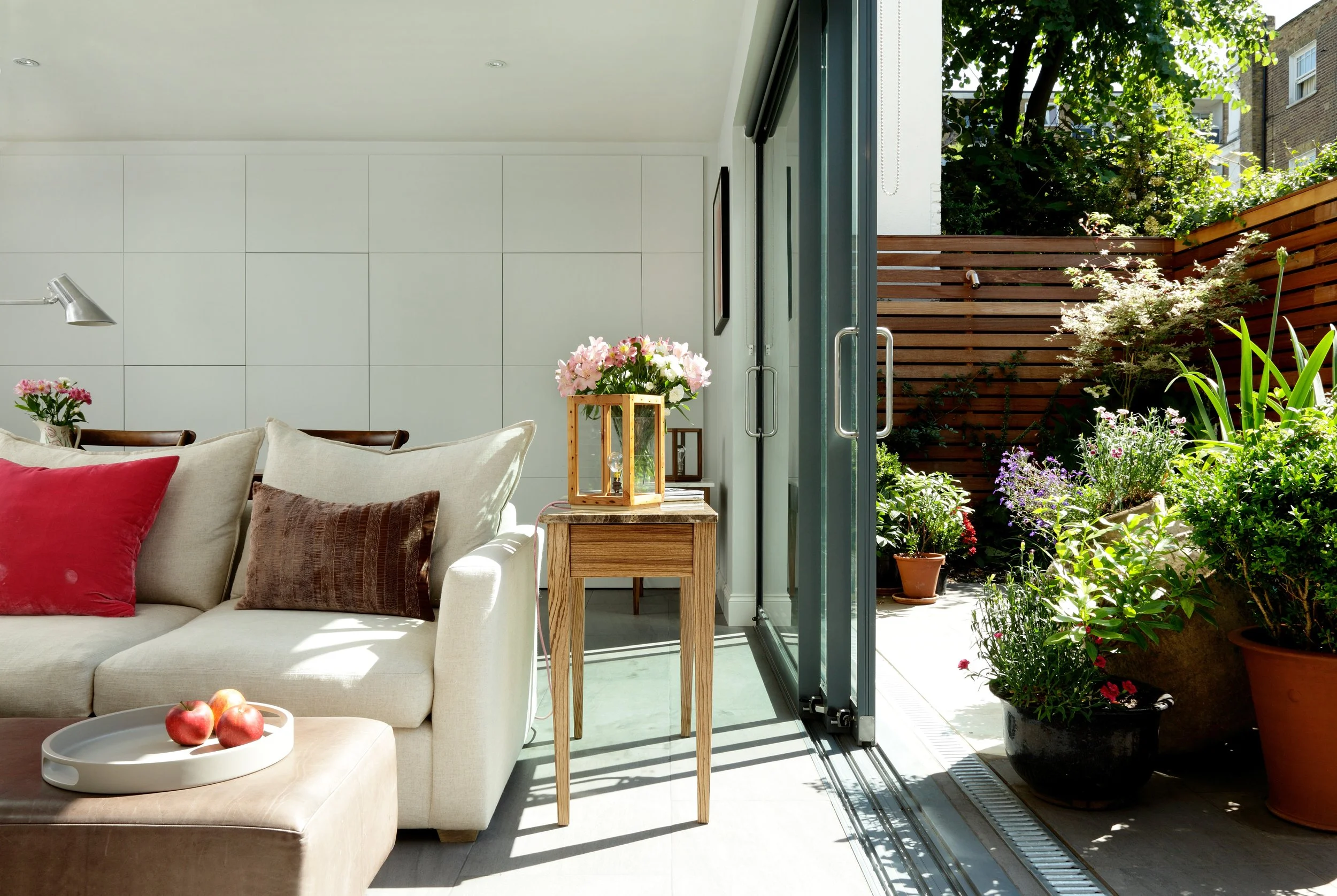 Living room with beige sofa, decorative pillows, side table with pink flowers, and open sliding glass door leading to a patio with potted plants and wooden fence, sunny weather.