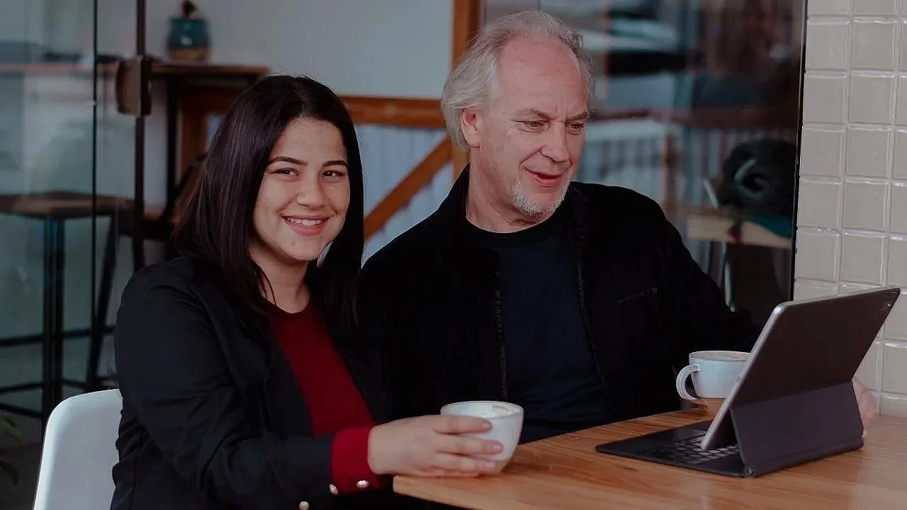 A young woman and an older man sitting at a wooden table in a cafe, smiling at the camera while holding cups of coffee. The man is looking at a tablet on the table.