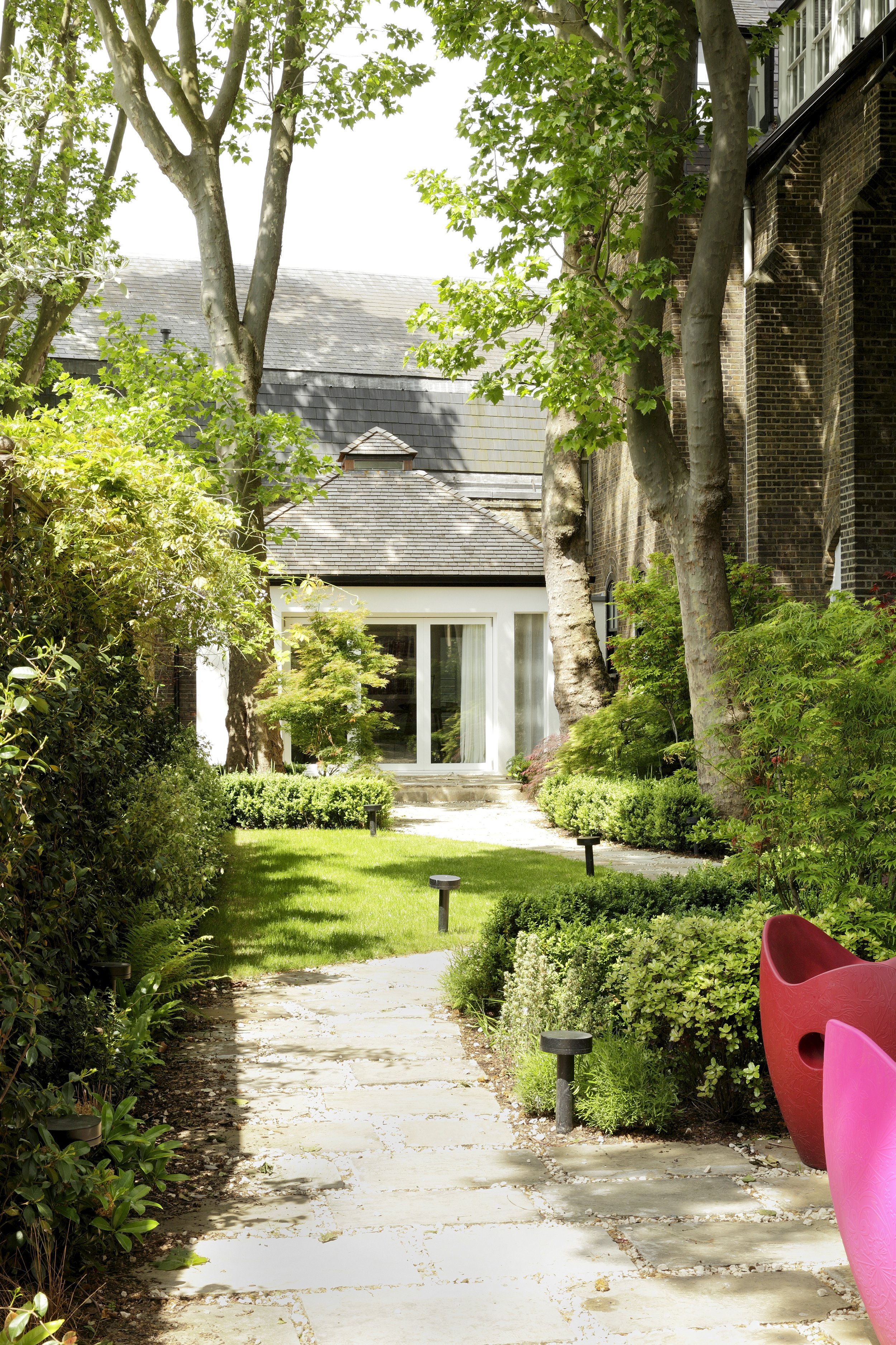 A lush garden pathway leading to a modern house with large glass doors, surrounded by trees, bushes, and green grass, with a pink and red outdoor chair on the right.