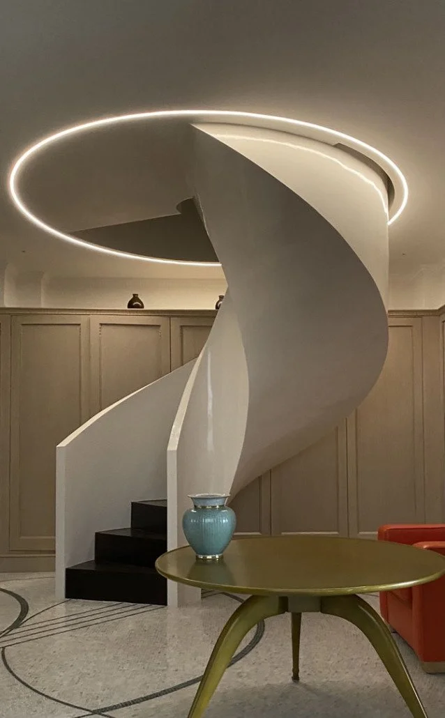 Interior of a modern space with a white spiral staircase, a round ceiling light, a green table with a blue vase, and beige paneled walls.