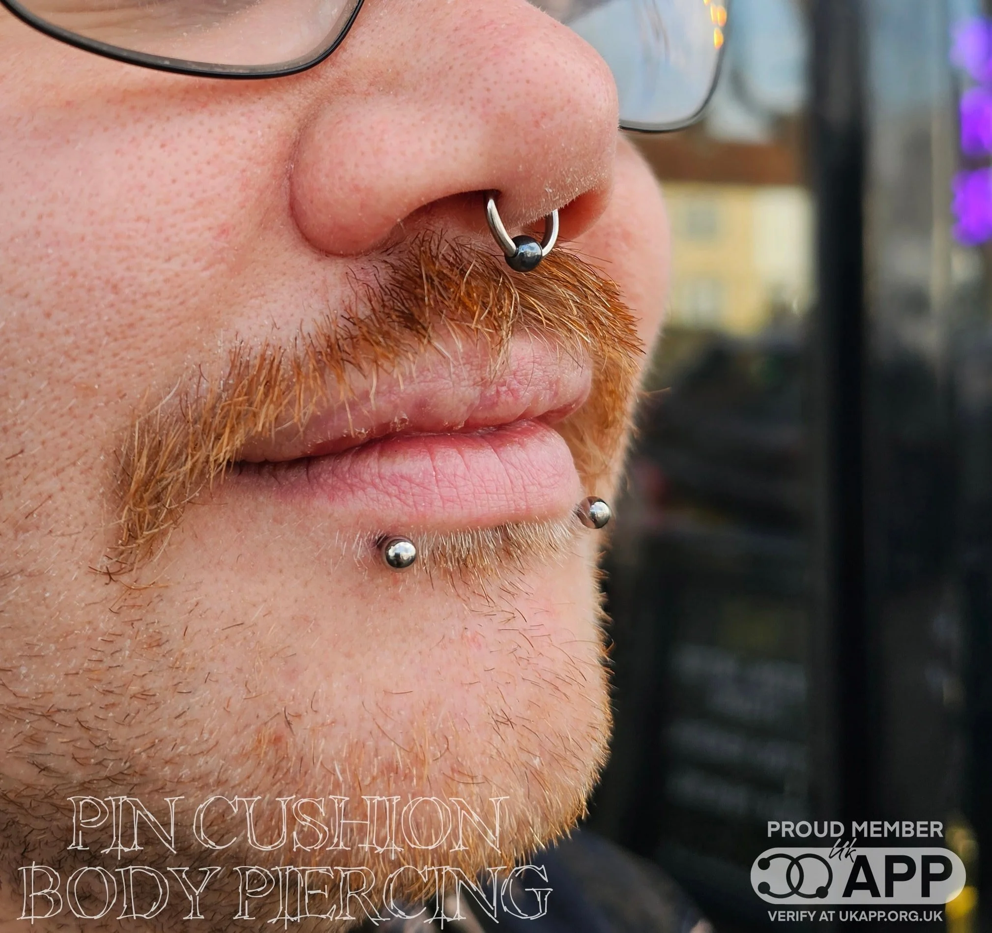Close-up of a person's face with glasses, multiple facial piercings including a septum ring and lip piercings.