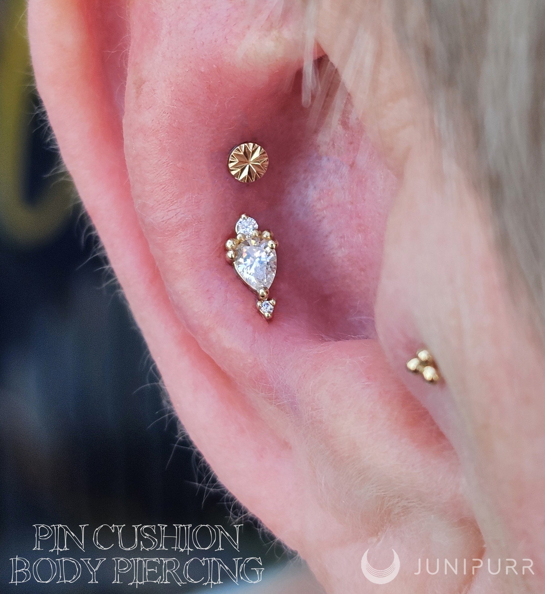 Close-up of a person's ear with multiple piercings, including a gold circular stud and a cluster of diamond-like earrings. The background is black with white text, partially visible, that reads 'PIN CUSHION' and 'BODY PIERCING.' The logo and name 'JUNIPURR' are in the lower right corner.