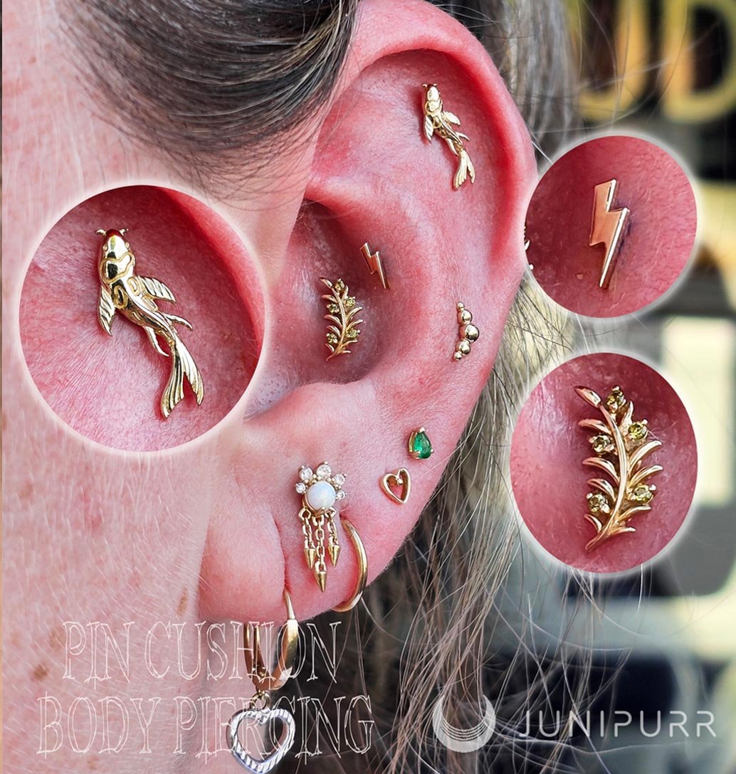 Close-up of a person’s ear adorned with multiple gold earrings including a fish-shaped earring, a leaf-shaped earring, a lightning bolt ear cuff, a star-shaped earring, a small round ball earring, a small heart-shaped earring, a green gemstone earring, and a dangling earring with a pearl and gold accents.