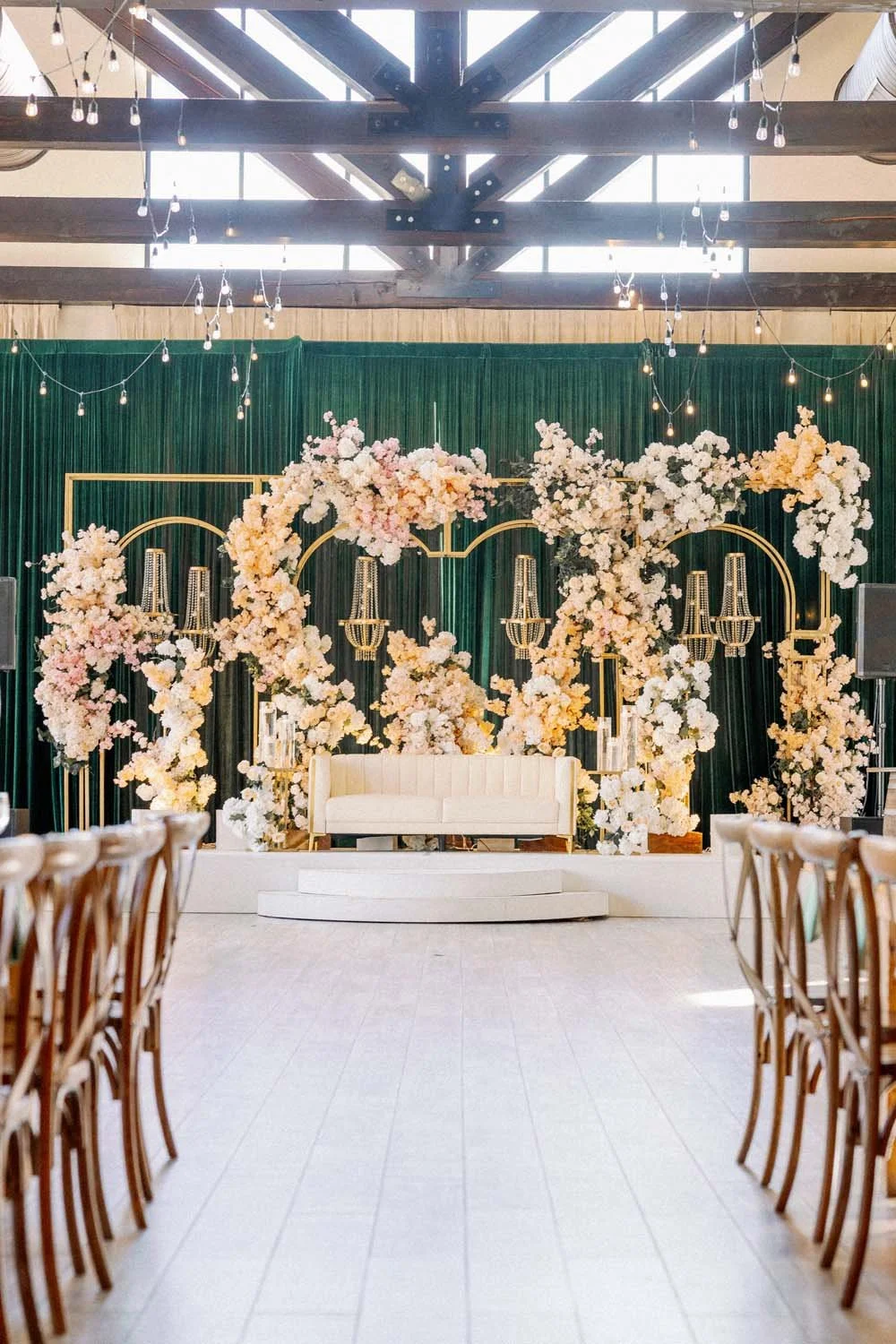 Grand reception stage inside The Barn featuring a deep emerald drape backdrop, crystal chandeliers, and asymmetric blush floral arches