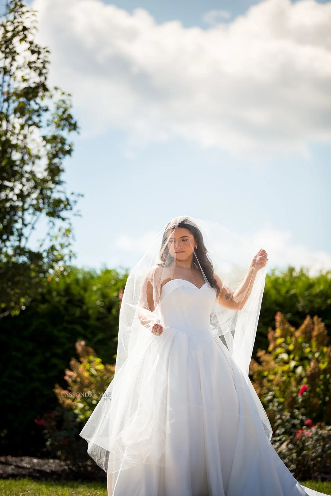 Bride in a white strapless wedding gown holding up her sheer cathedral veil under a bright blue summer sky in New York