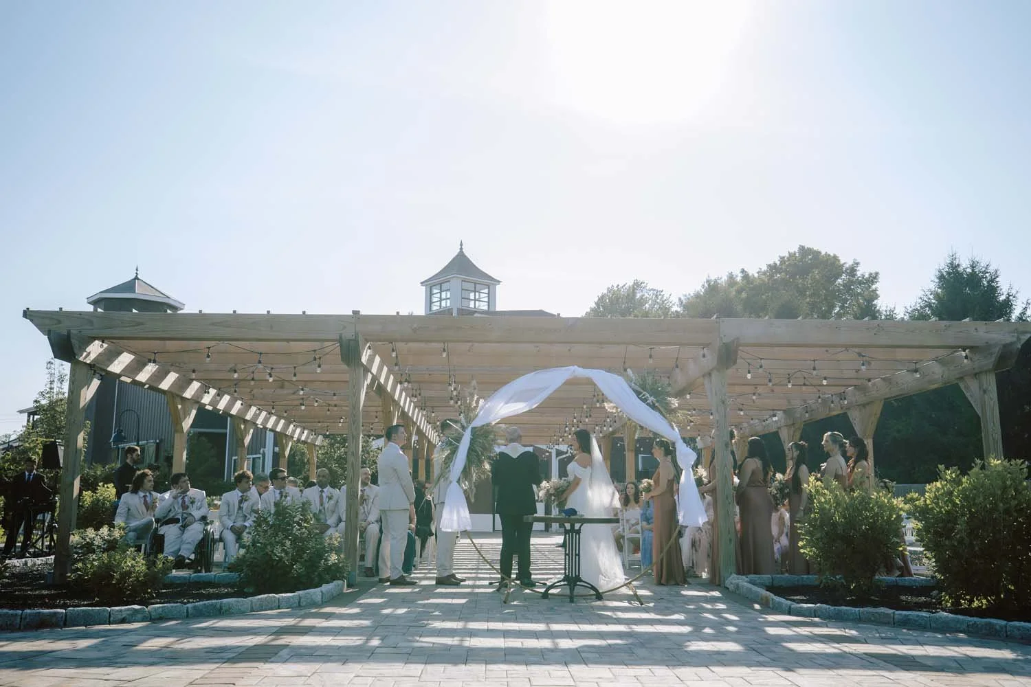 Jasmine and Brett exchanging vows under the sunlit wooden pergola at The Barn at Villa Venezia