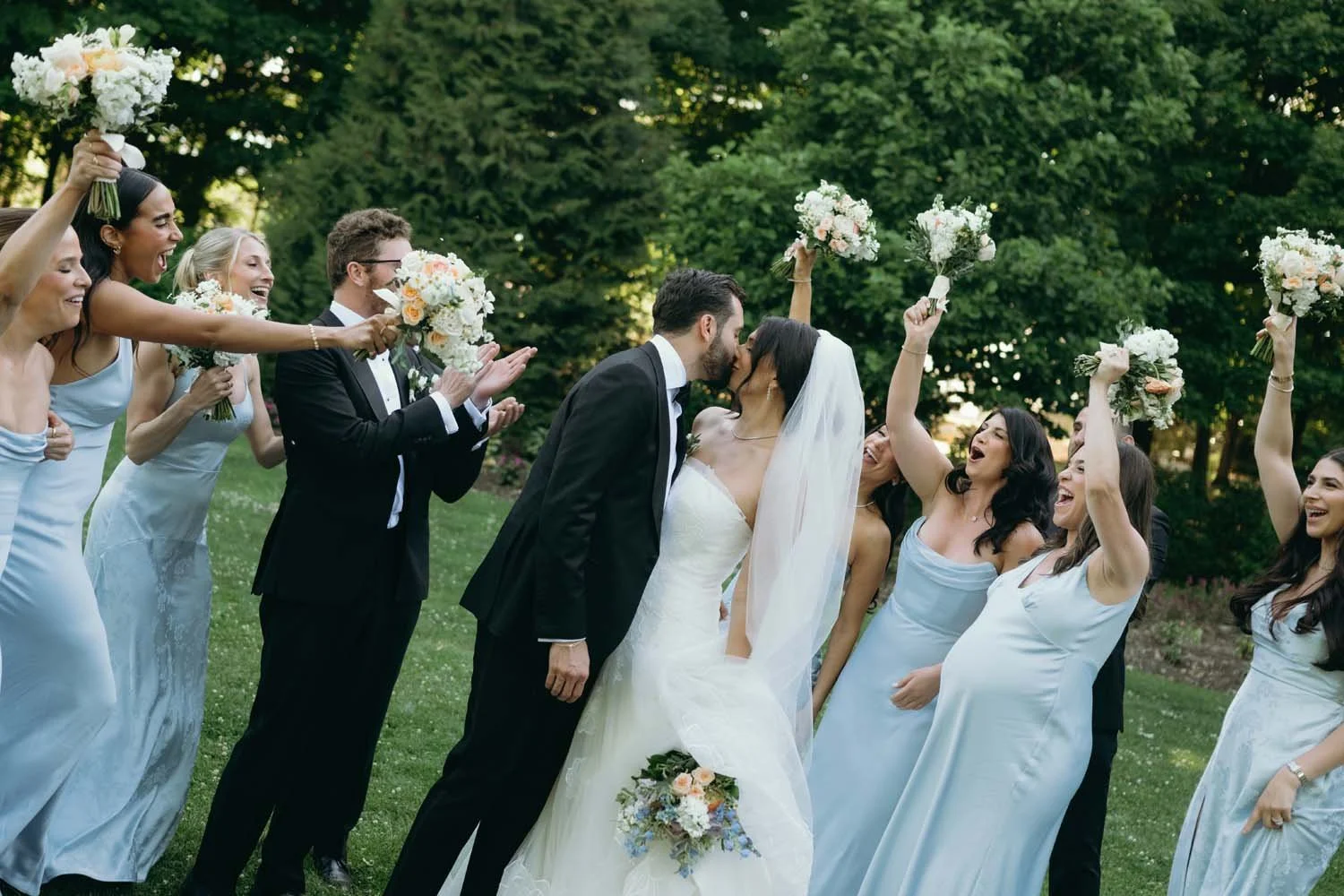 Bride and groom kissing outdoors surrounded by cheer from bridesmaids in baby blue dresses and groomsmen in tuxedos