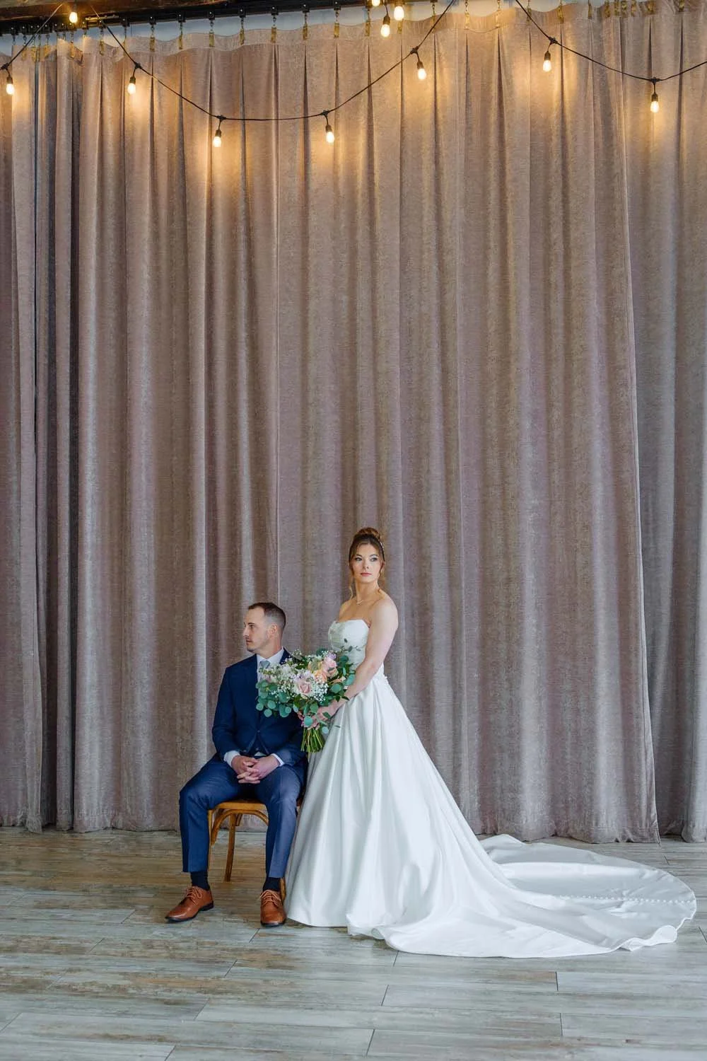 Bride and groom posing inside the Farmview Room ballroom beneath string bistro lights and grand cascading drapes.