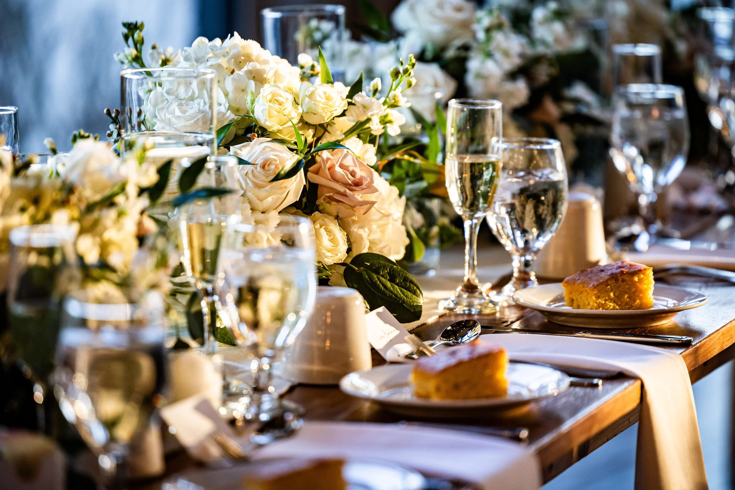 Warm, sunlit detail shot of a wedding reception table at The Barn at Villa Venezia with champagne flutes, white floral arrangements, and golden cornbread starters.
