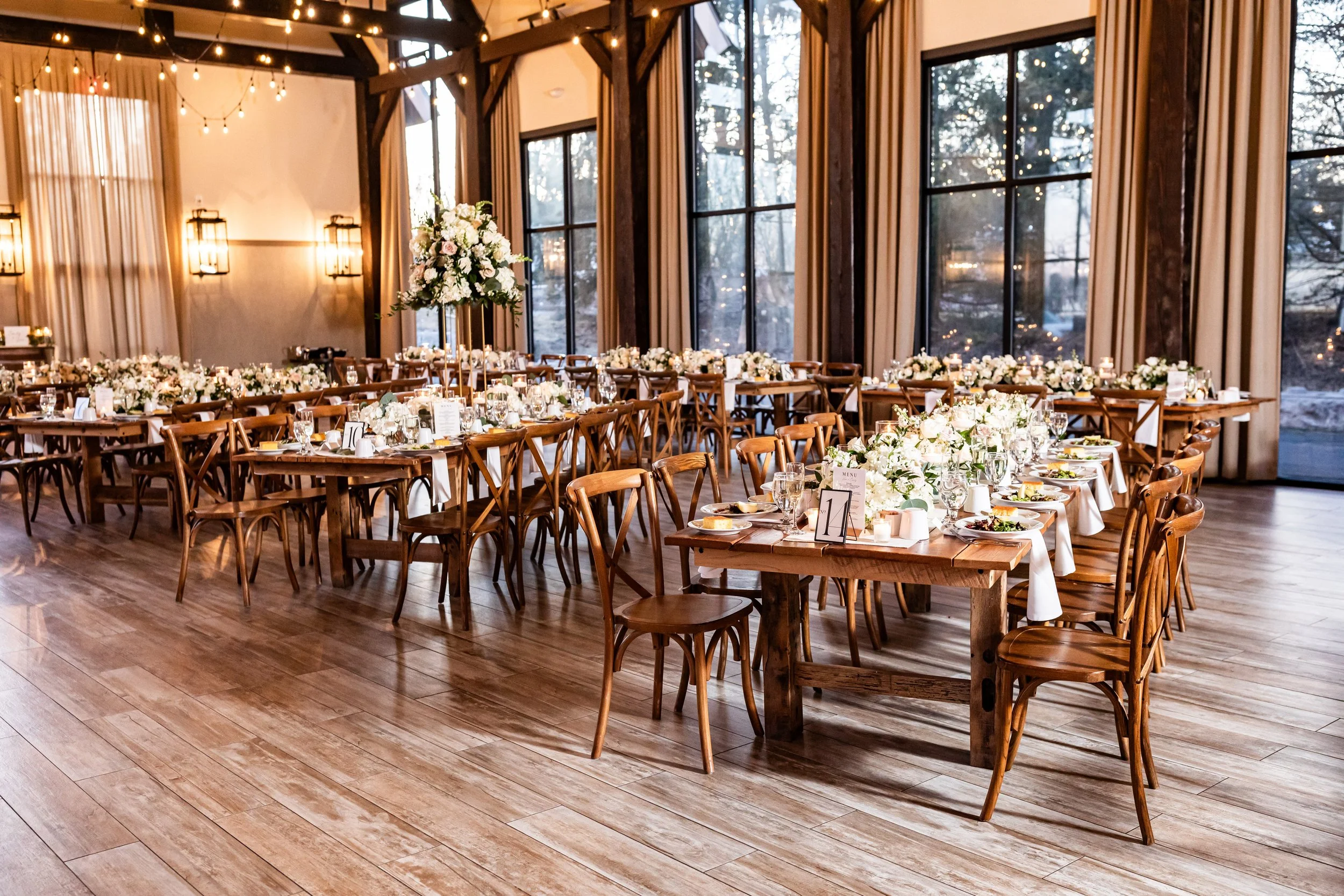 Wide shot of the indoor reception space at The Barn at Villa Venezia, highlighting the expansive windows, high ceilings, string lighting, and organized wooden wedding tables.