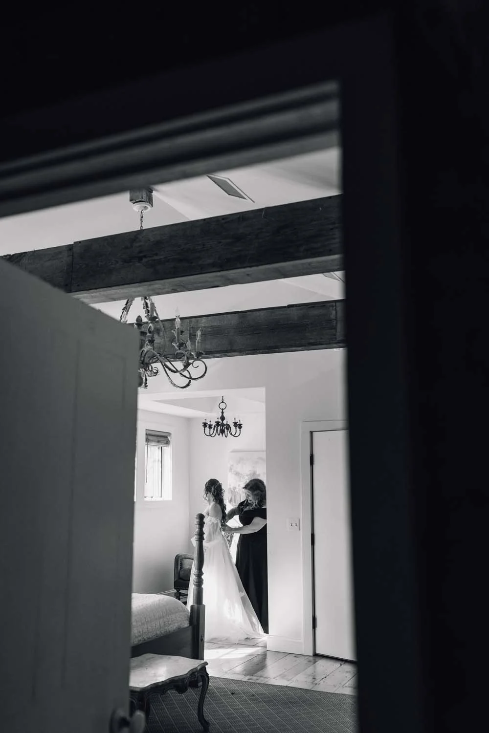 Black and white photo of a mother helping her daughter step into her wedding dress under a rustic wooden beam