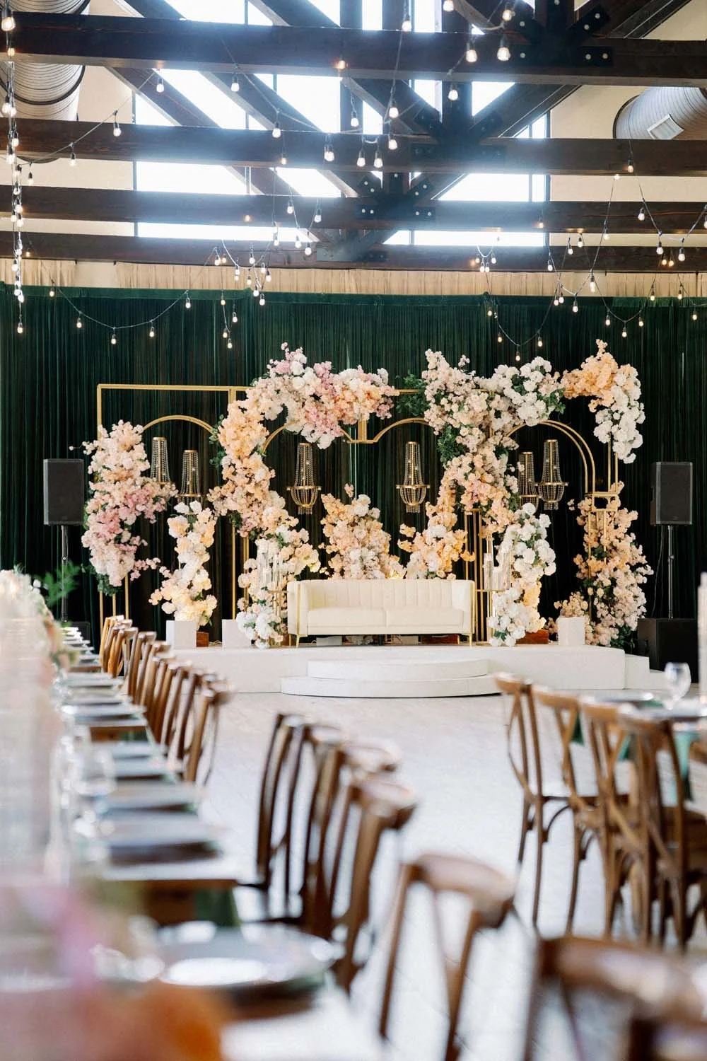 Wide indoor shot of long wooden reception tables and cross-back chairs leading toward a grand lit stage