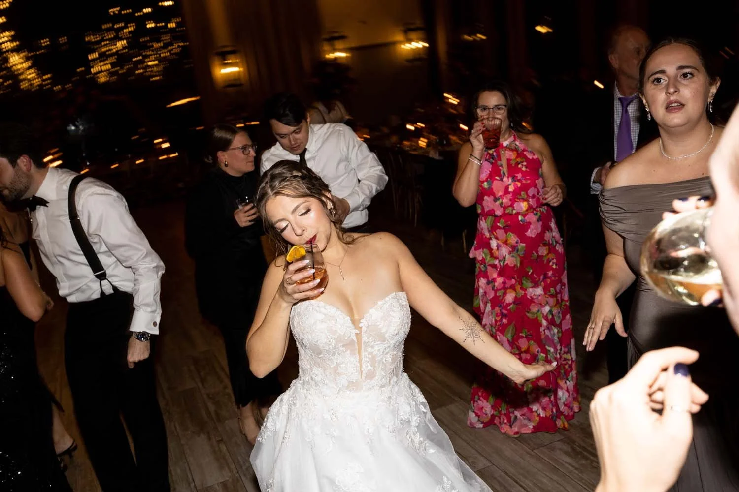 A bride sips on her drink while dancing with her wedding party and guests