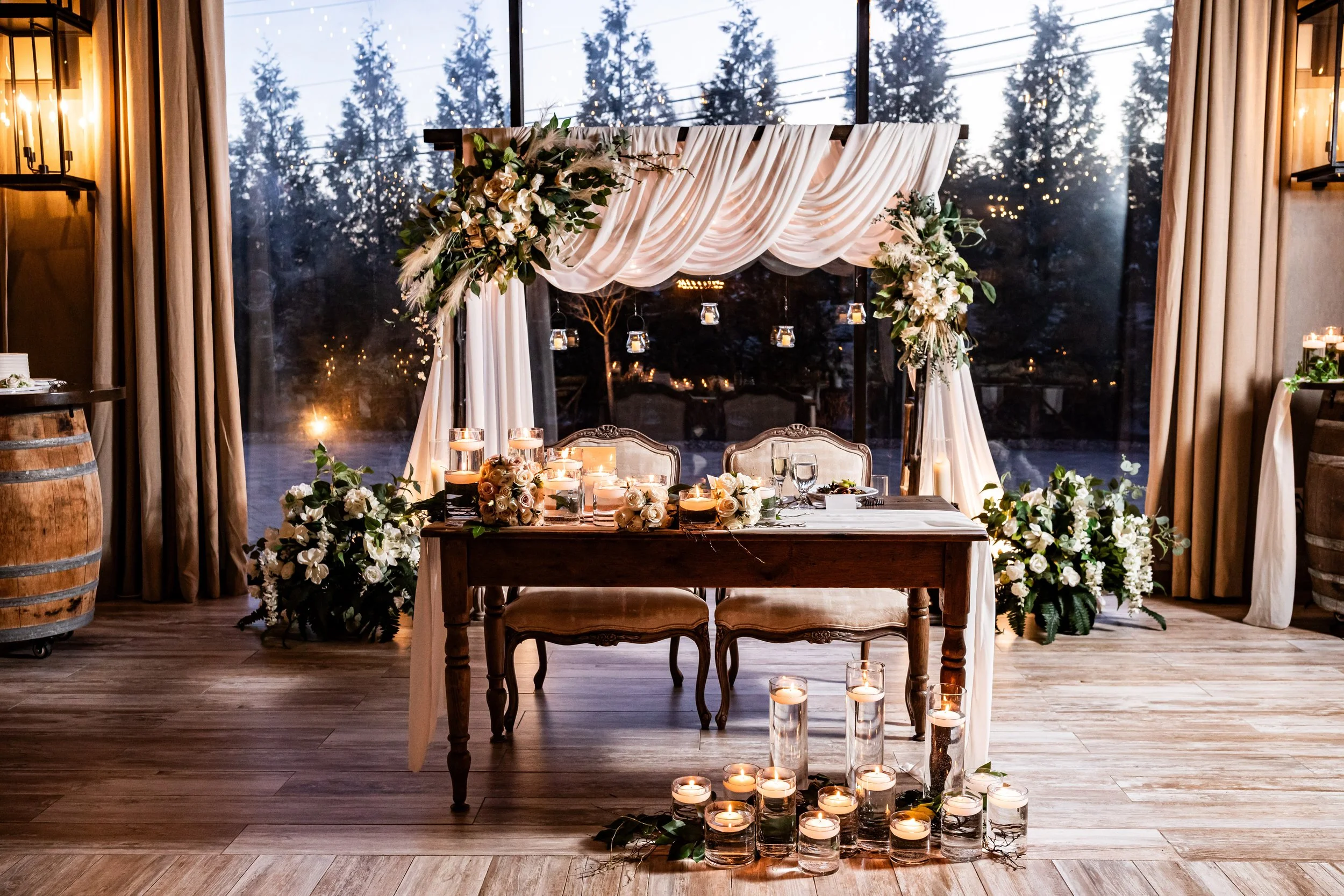 Elegant wedding sweetheart table at The Barn at Villa Venezia, decorated with draped white fabric arch, cascading white and green florals, and numerous floating candles in glass cylinders.