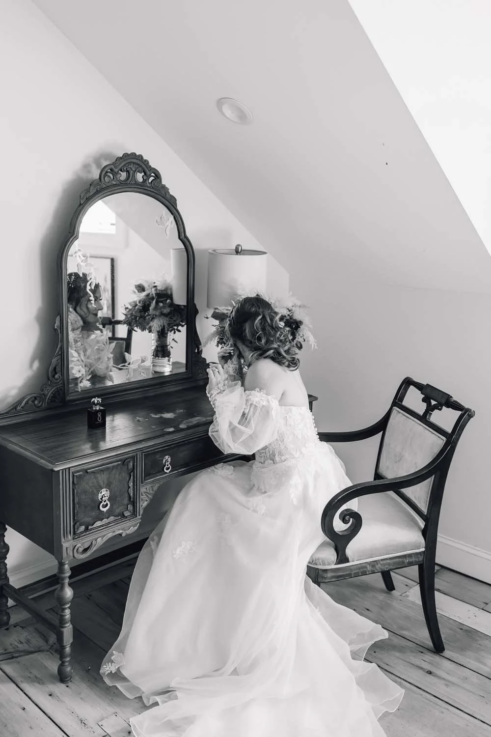 A black and white image of a bride sitting at a vintage vanity putting on earrings