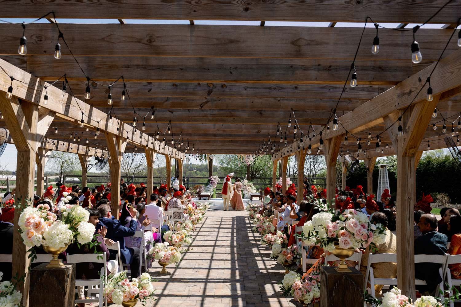 Beautiful outdoor wedding ceremony under a wooden pergola with string bistro lights and gold floral urns lining the aisle.