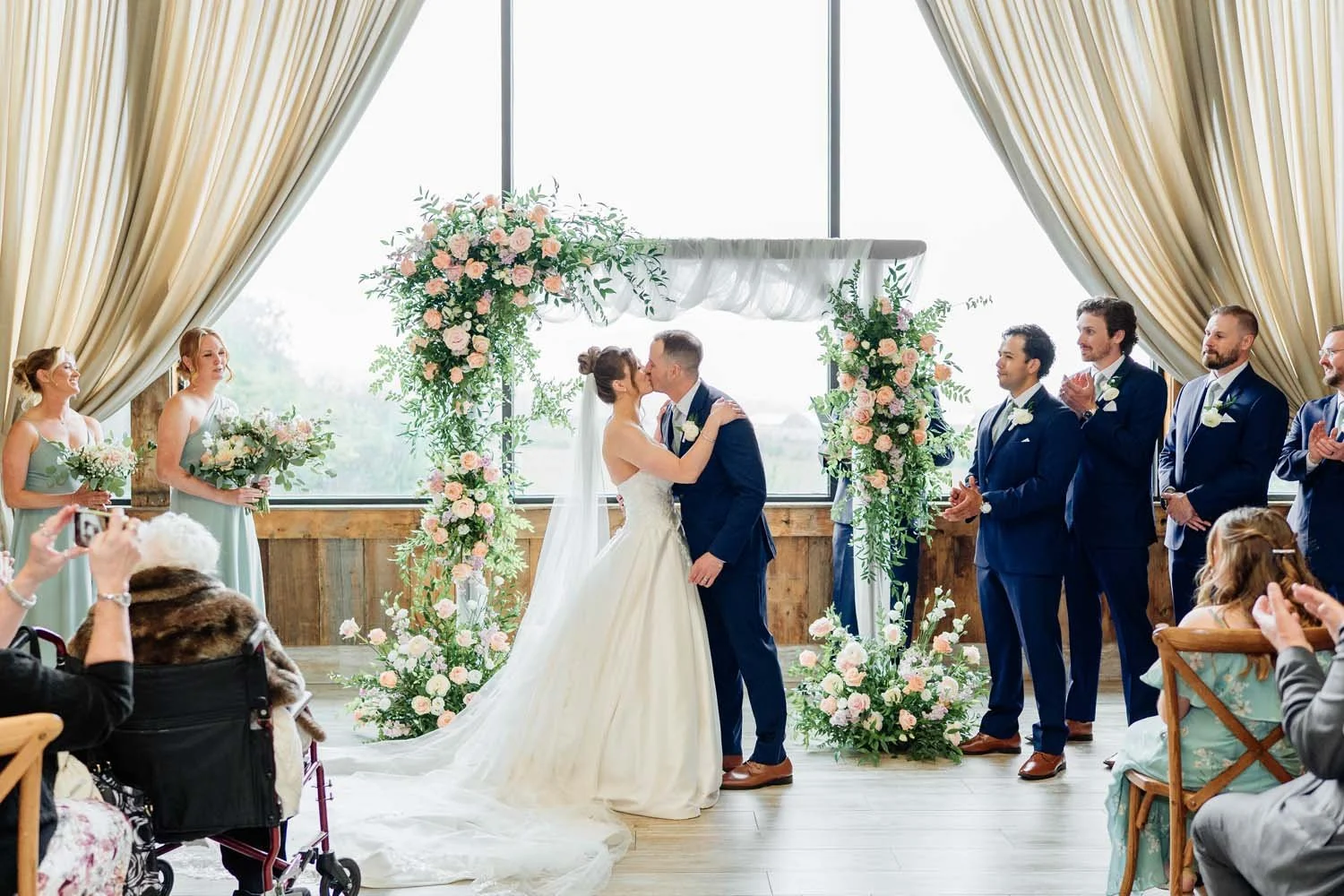 Bride and groom sharing their first kiss at an indoor ceremony in the Farmview Room in front of a floral arch and wedding party.