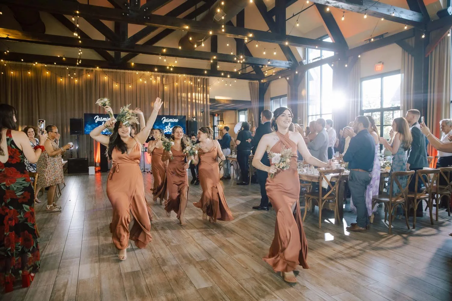 Bridesmaids in copper gowns dancing excitedly into the reception hall at The Barn at Villa Venezia