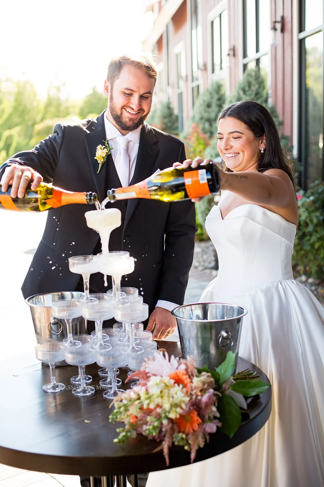 Bride and groom smiling while pouring bottles of champagne into a glass tower outside at The Barn at Villa Venezia