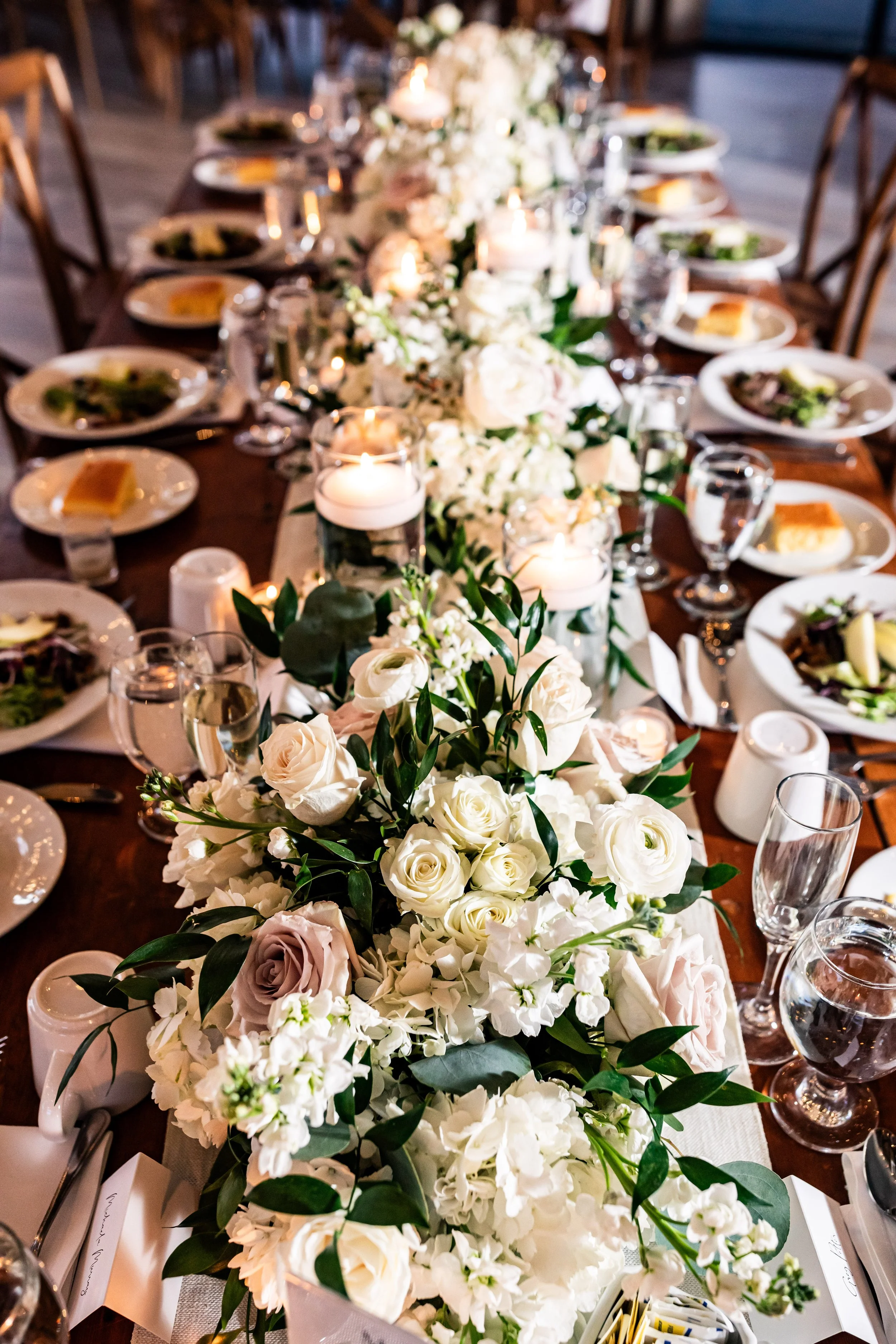 Overhead view of a long farm table wedding reception setup featuring a lush white and dusty rose floral runner, floating candles, and plated salads
