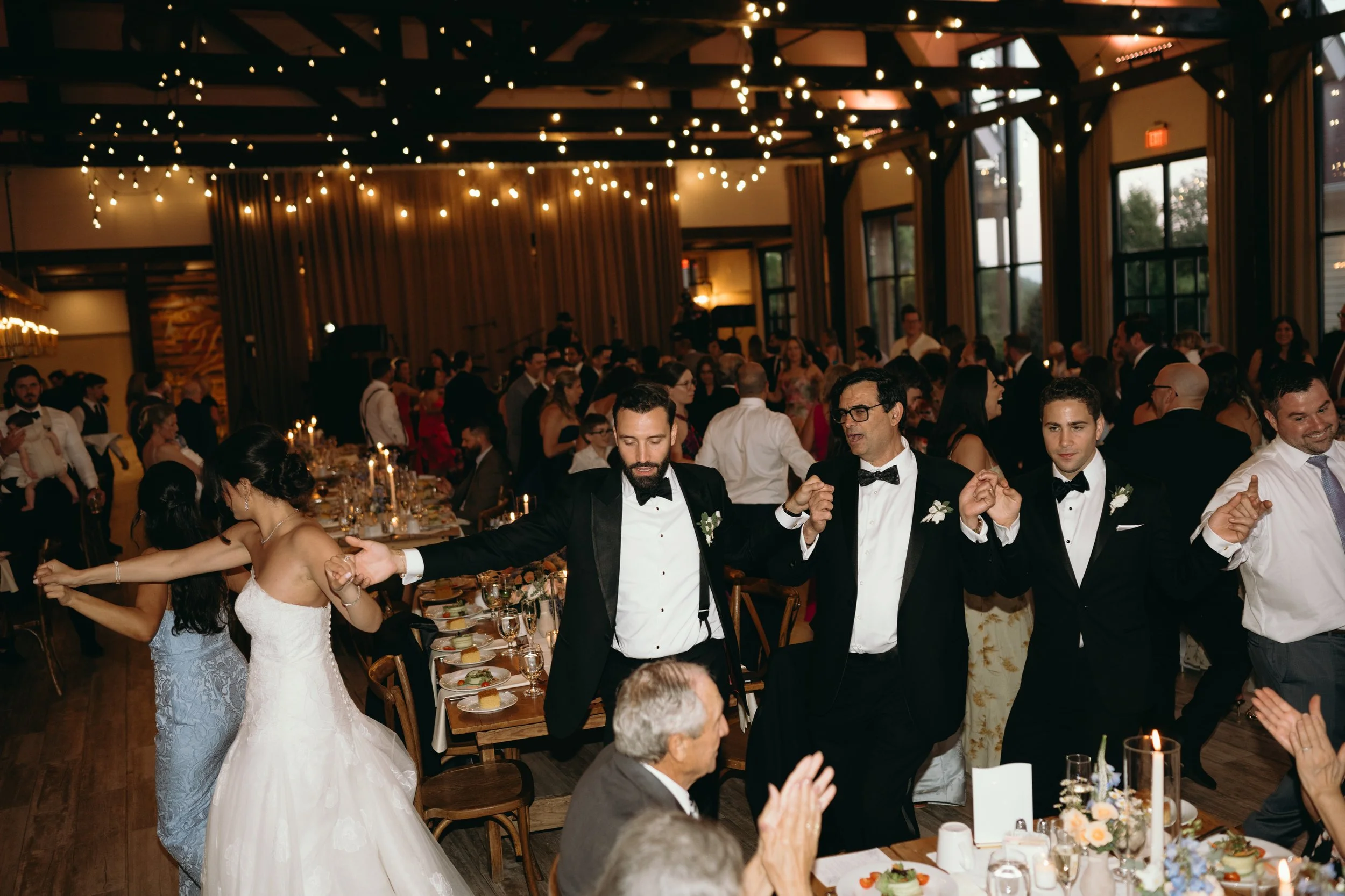 Bride in strapless gown and groom in tuxedo dancing with guests under bistro lights at The Barn at Villa Venezia